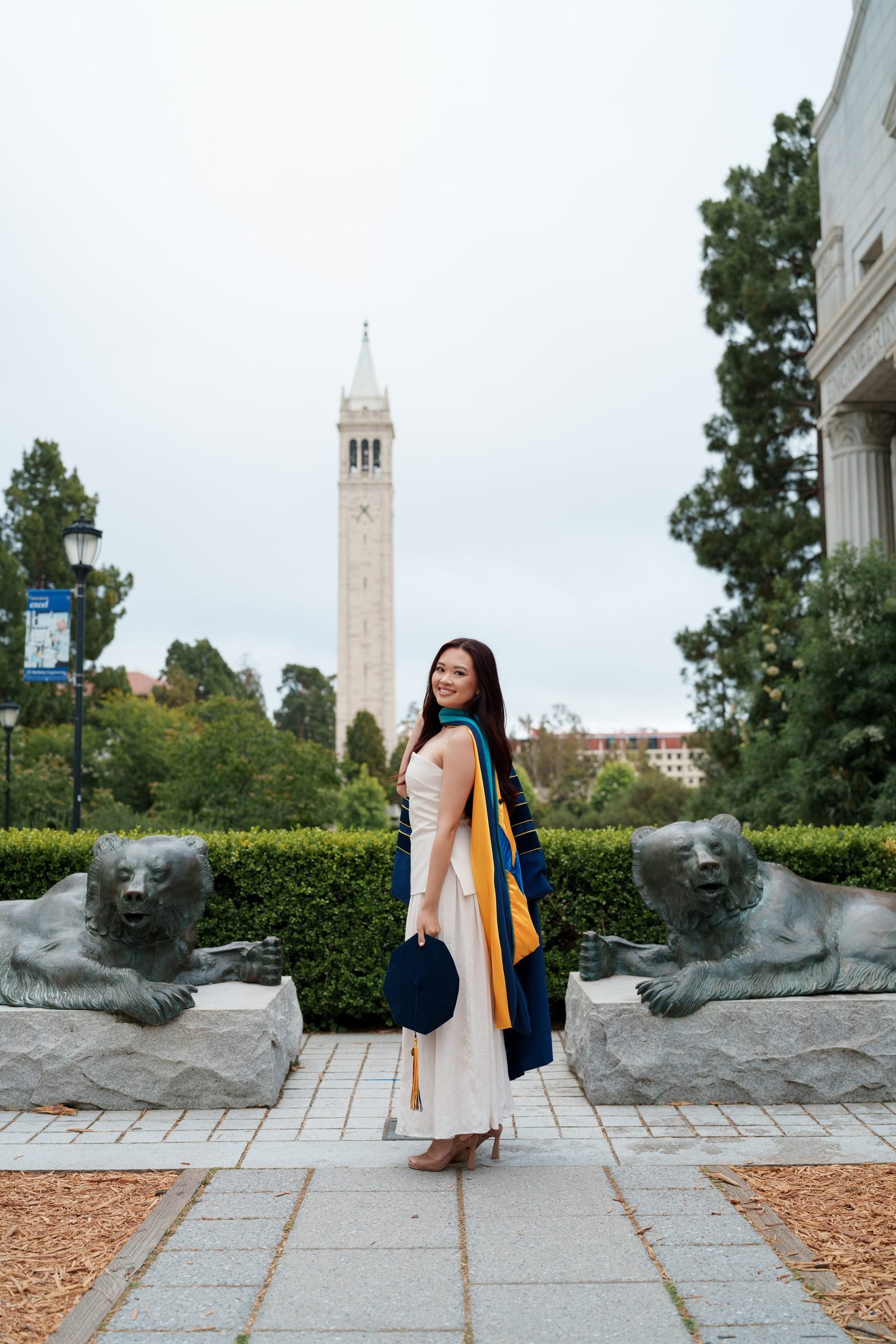 Trang stands between two bronze bear sculptures near Doe Library, glancing back over her shoulder with a warm smile. She holds her navy cap at her side, doctoral regalia draped over a white dress, with Sather Tower rising behind her under soft overcast light.