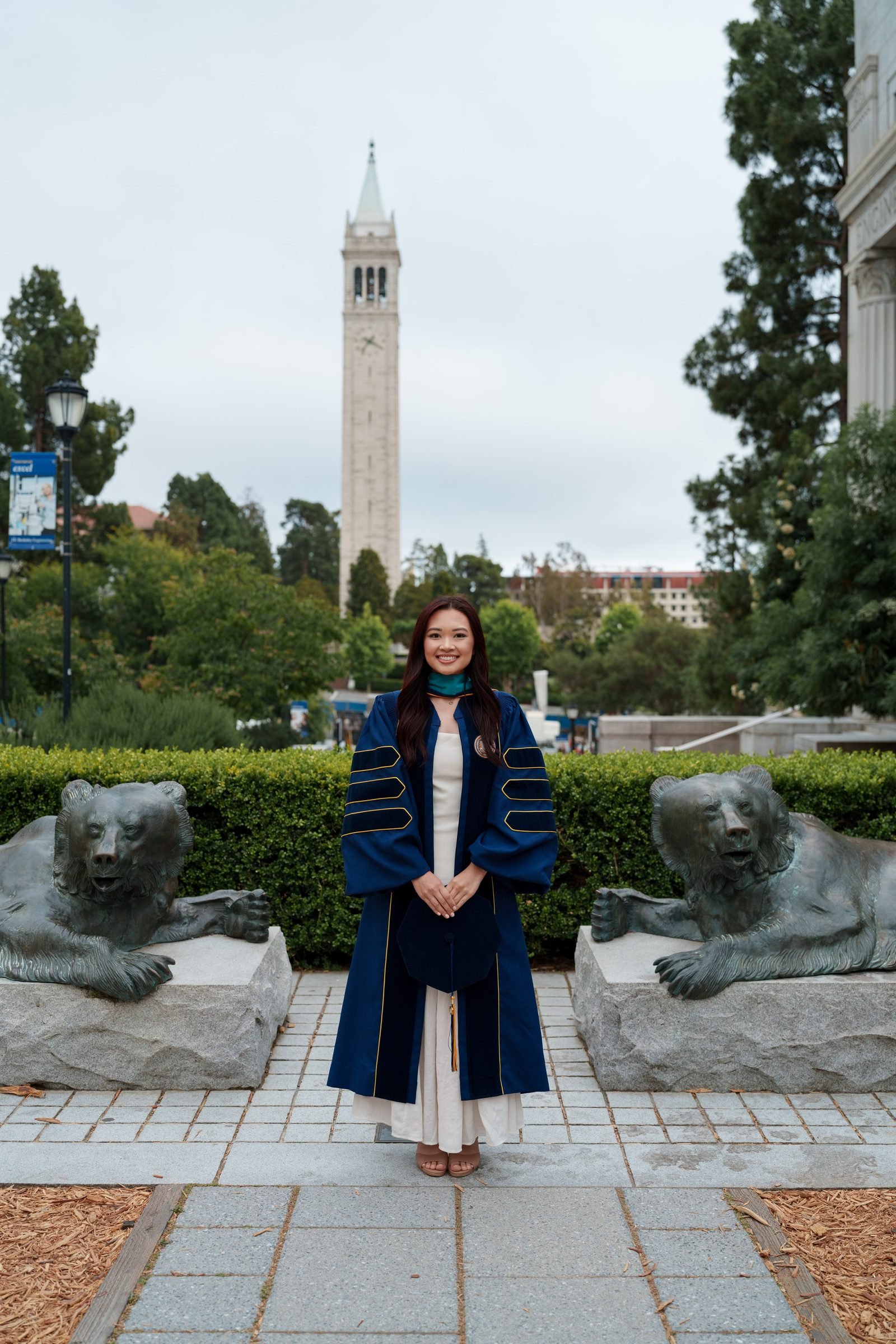 Trang stands centered between two bronze bear sculptures with the Campanile rising behind her. She wears a navy doctoral robe with gold trim over a cream dress, smiling warmly under soft overcast Bay Area light.