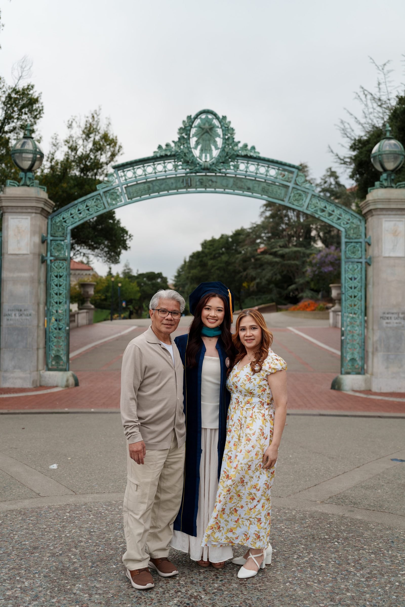 Graduate in navy cap and gown stands between her parents under Sather Gate on an overcast day, all three smiling warmly at the camera against the iconic green iron archway.