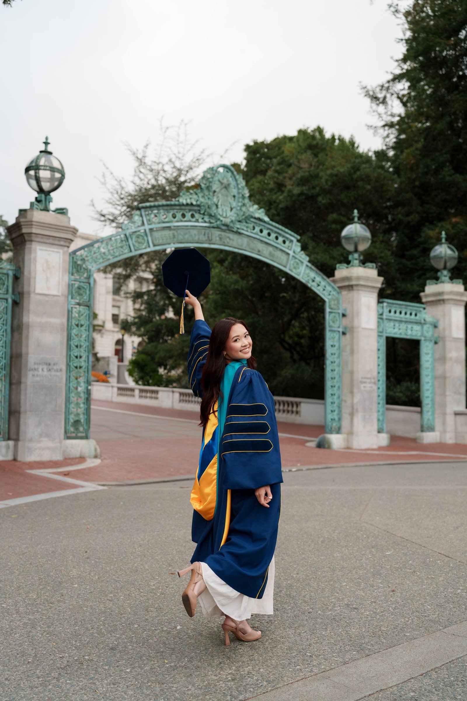 Trang walks through Sather Gate in a navy doctoral gown with gold trim and teal hood, cap raised overhead, glancing back with a wide smile on an overcast morning