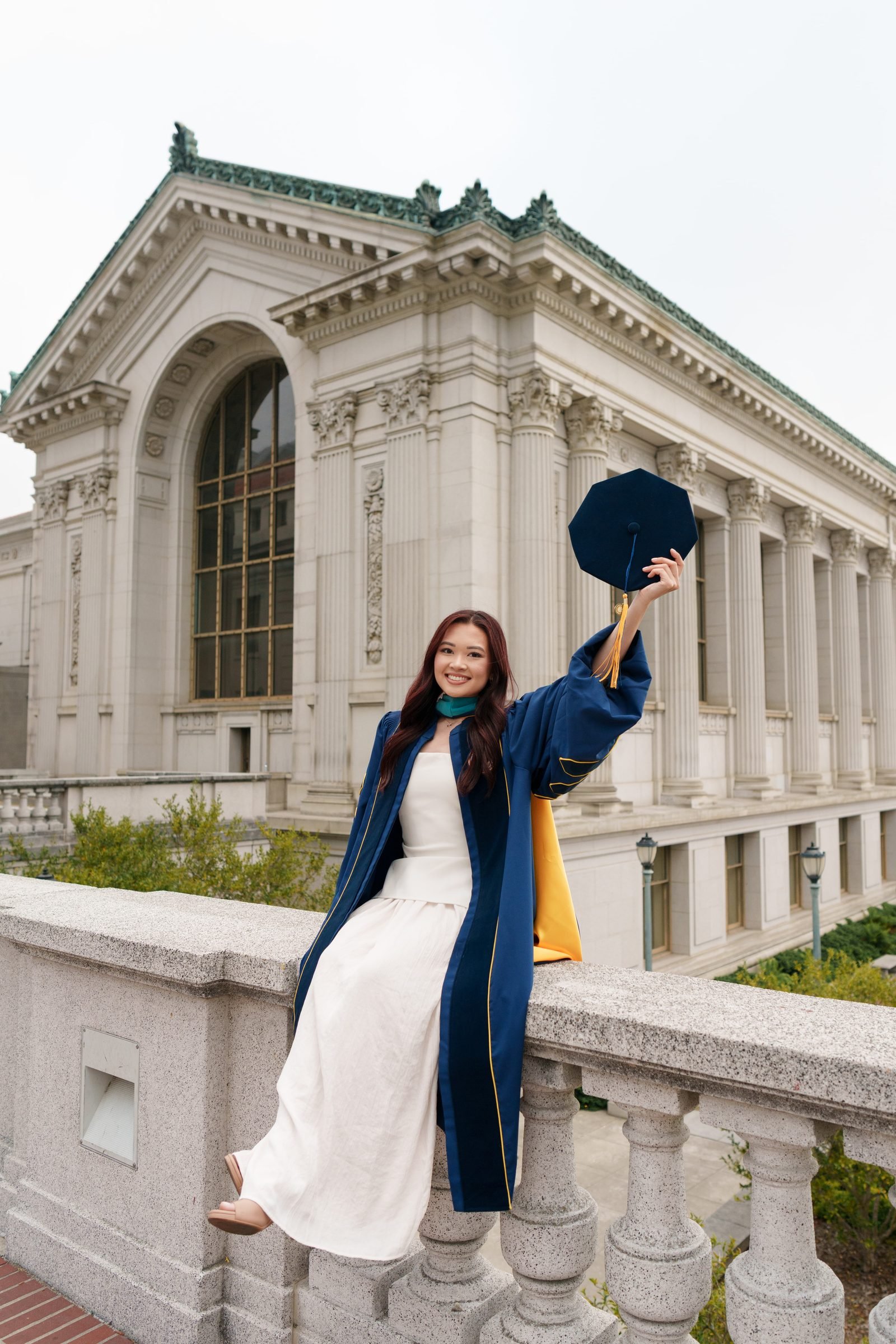 Trang sits perched on a stone balustrade in front of a grand neoclassical building, wearing a navy doctoral gown over a white dress, holding her cap aloft and smiling at the camera under soft overcast light.