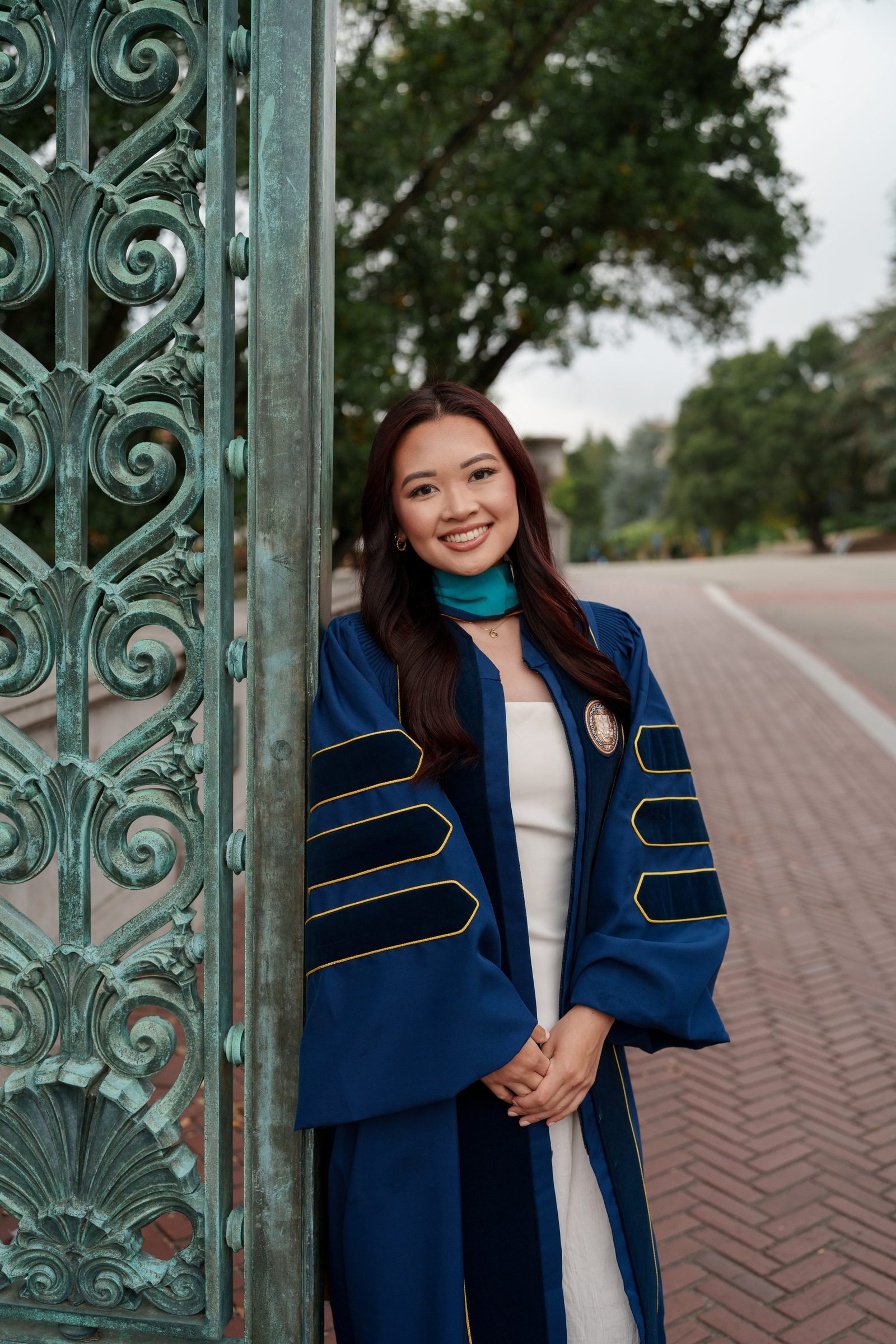 Trang leans against the ornate verdigris ironwork of Sather Gate, wearing a navy doctoral gown with gold and black velvet trim over a white dress, smiling warmly on a soft overcast day.