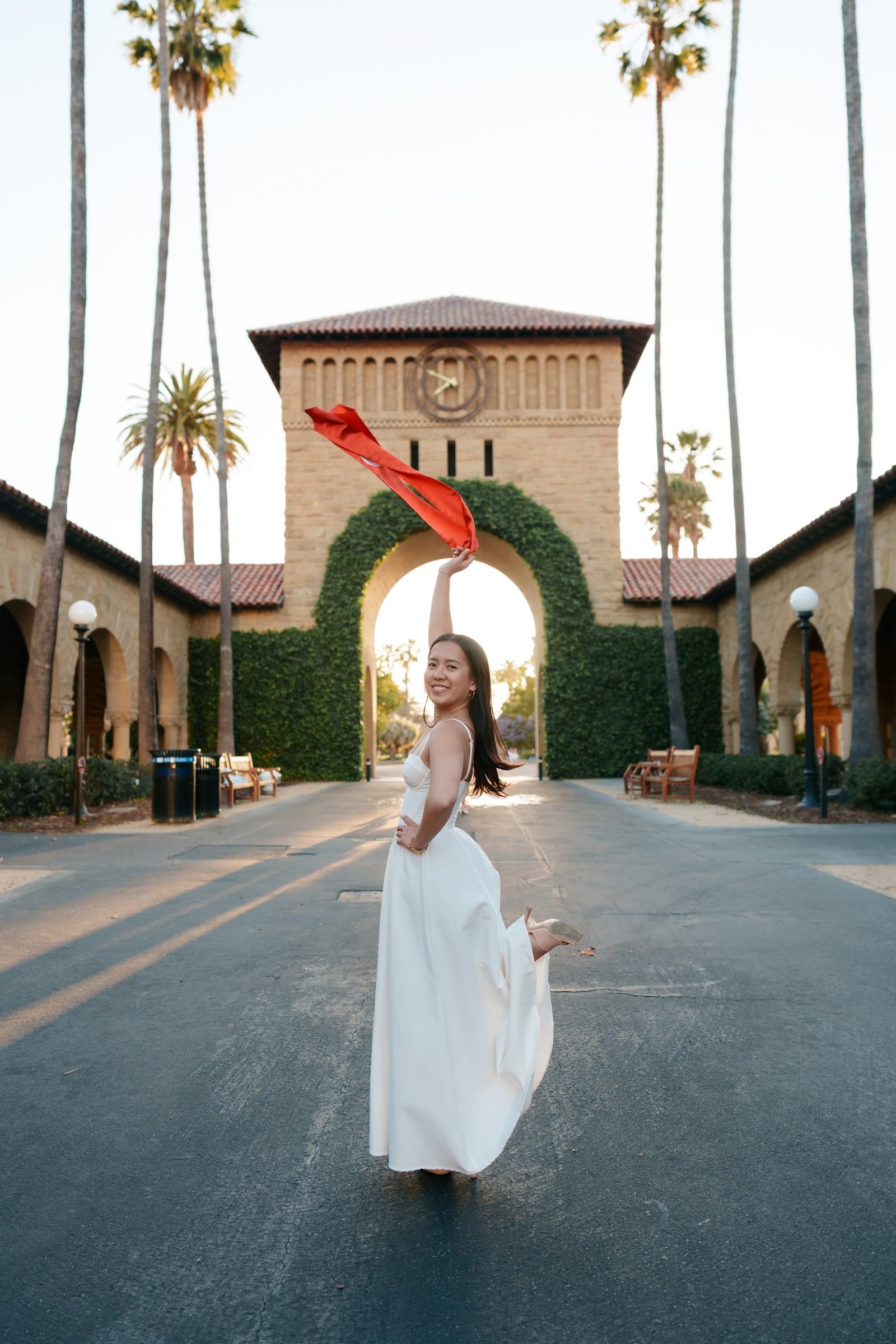 Chantal in a white maxi dress twirls her red graduation stole overhead on the palm-lined Main Quad road, the ivy-covered clock tower arch framing her against a soft evening sky.