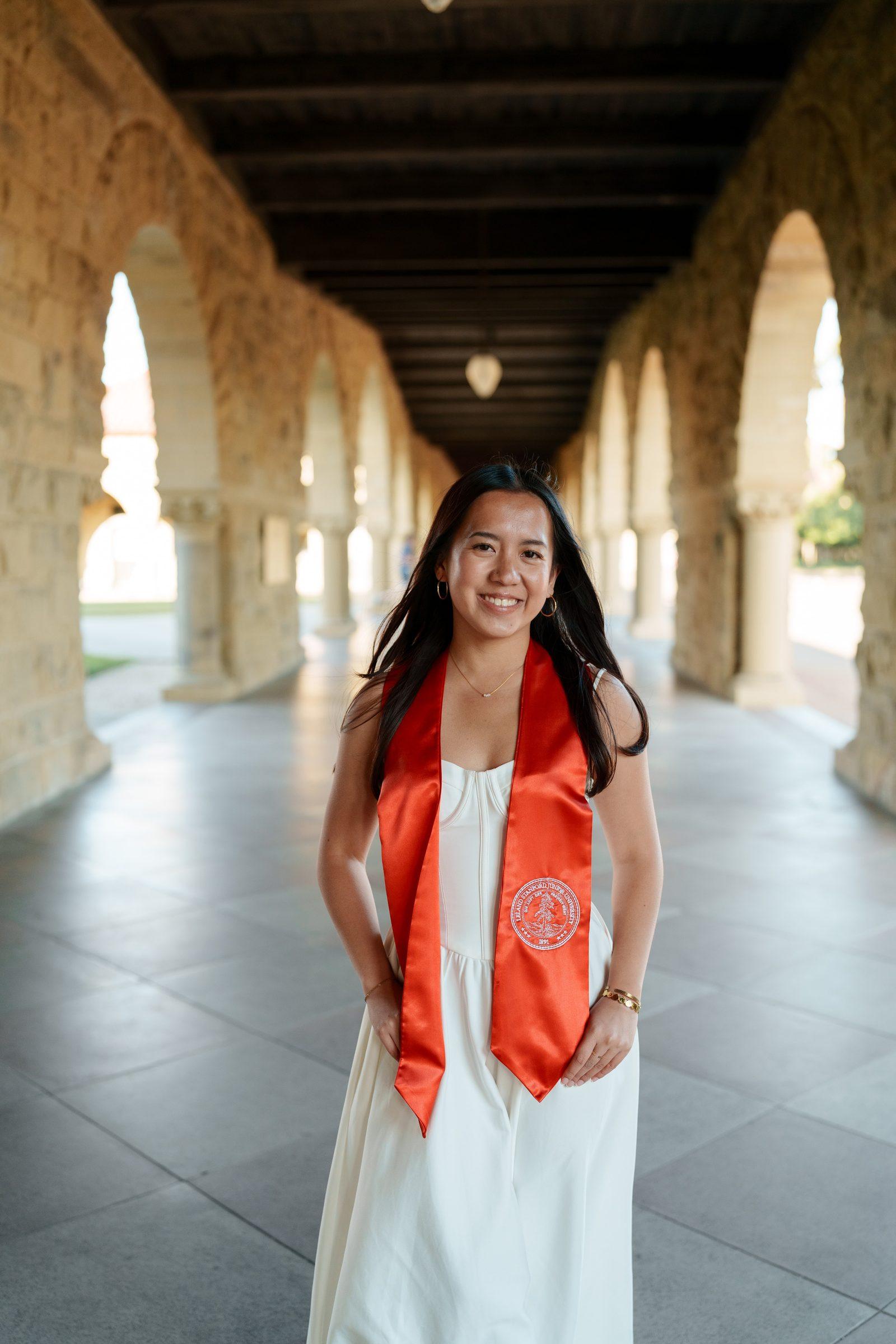 Chantal stands in Stanford's sandstone arcade corridor wearing a white dress and cardinal graduation stole, smiling directly at the camera with a long row of arches receding behind her.