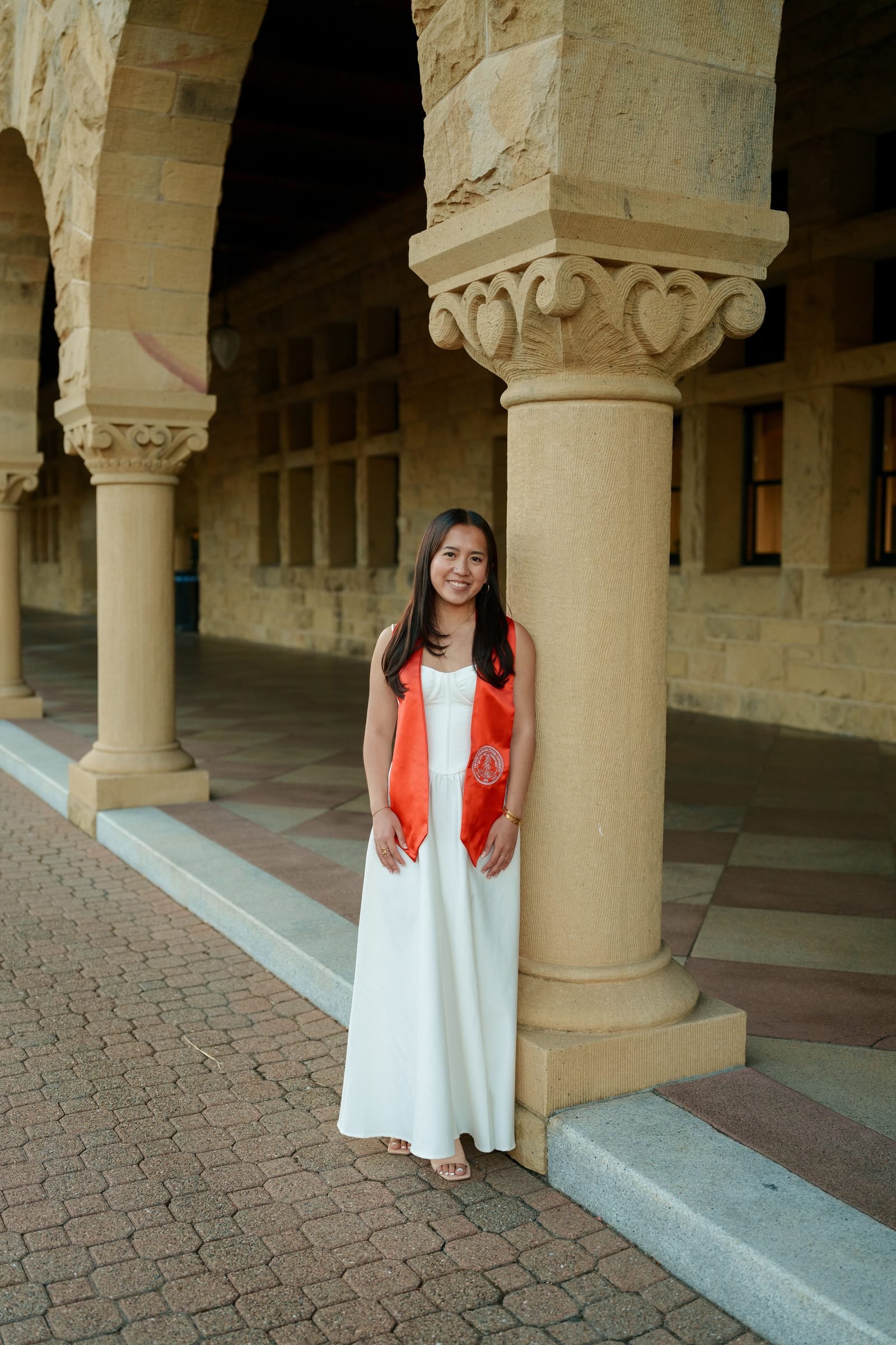Chantal leans against an ornately carved sandstone column in the Main Quad arcade, wearing a white dress and red graduation stole, smiling warmly in evening shade.