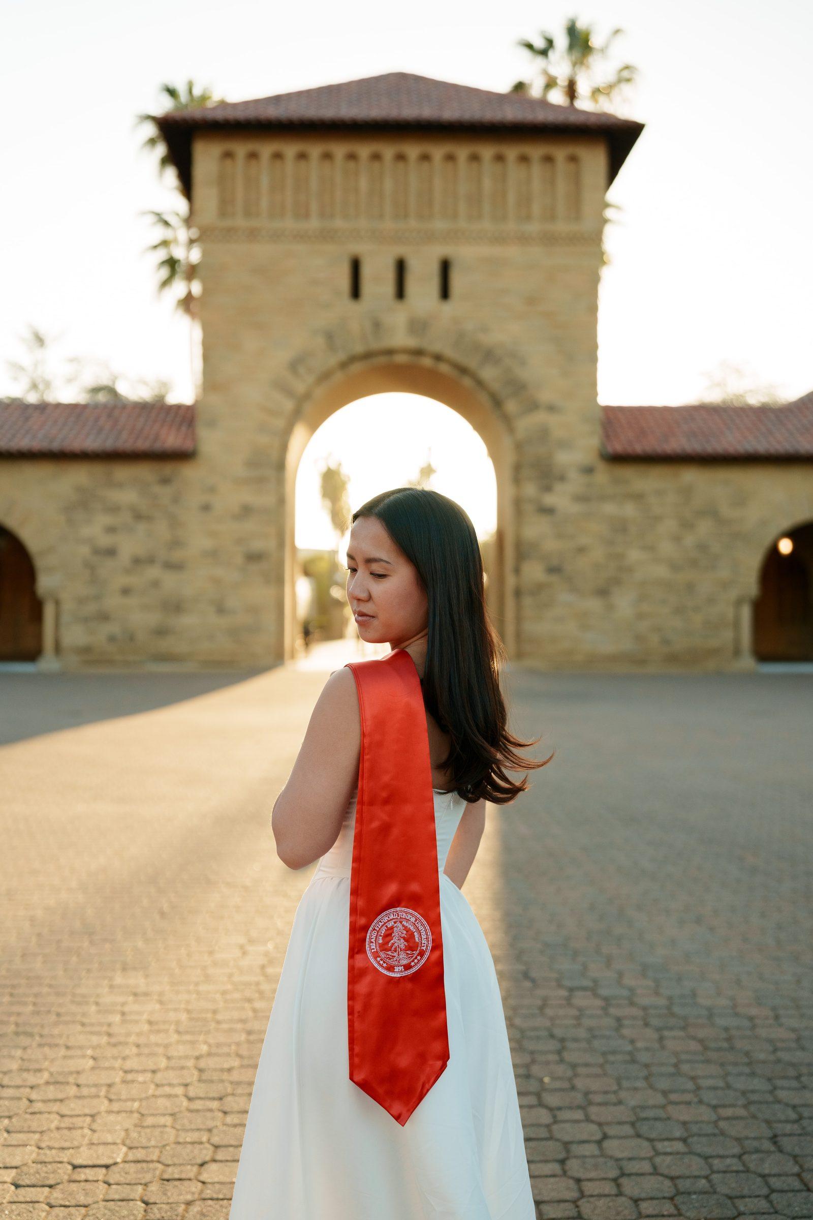 Chantal stands with her back to the camera in a white dress and red Stanford sash, looking over her shoulder toward the glowing Main Quad gateway arch with golden backlight at sunset.