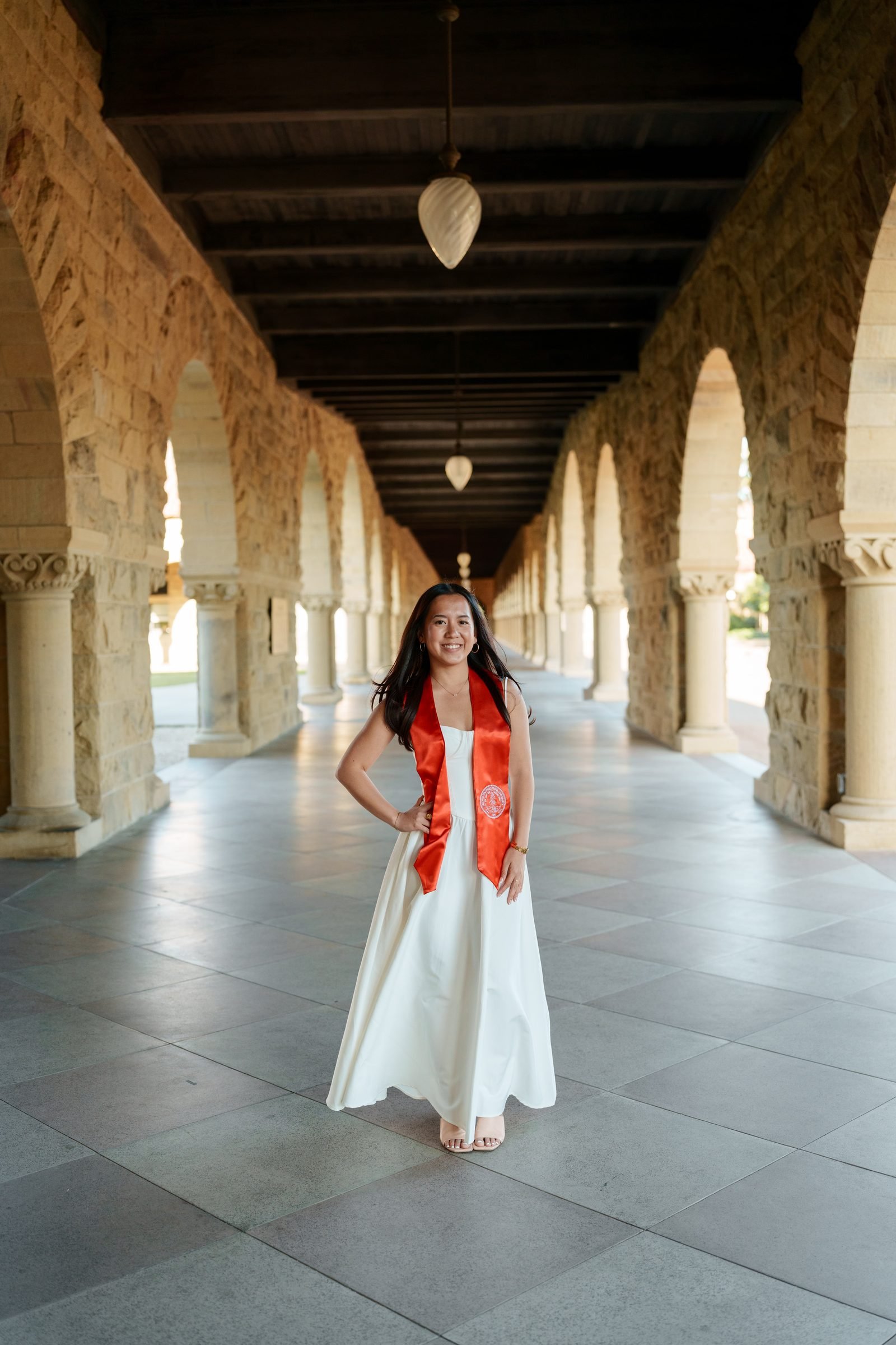 Chantal stands full-length in the Stanford Main Quad colonnade wearing a white maxi dress and cardinal red graduation sash, one hand on her hip, beaming confidently toward camera.