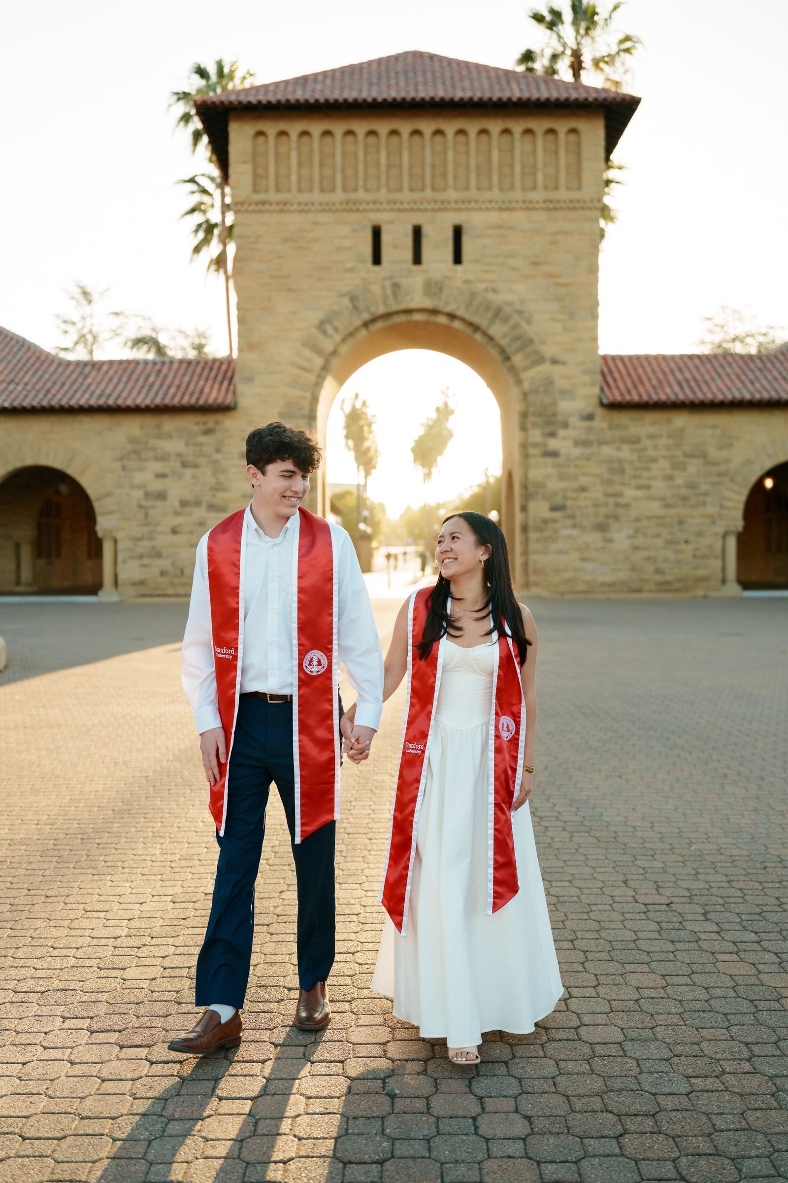 Two Stanford graduates in red sashes hold hands and walk through the Main Quad gateway at sunset, golden backlight pouring through the stone arch behind them as they smile at each other.