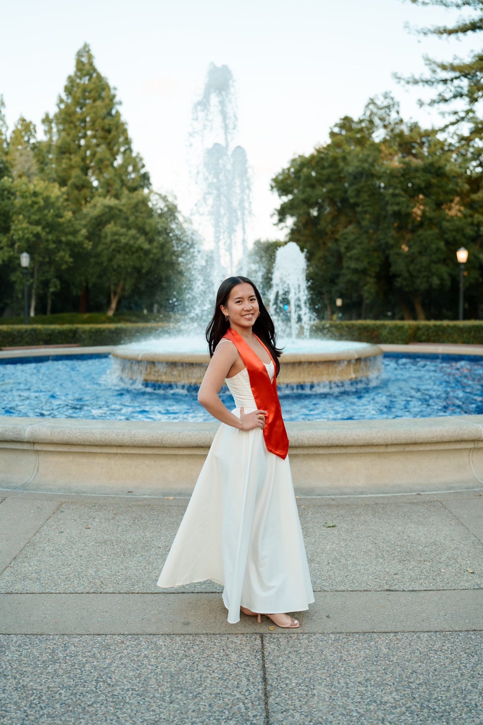 Graduate in a white floor-length gown and red satin stole stands confidently in front of a large campus fountain at dusk, hand on hip, smiling over her shoulder toward the camera