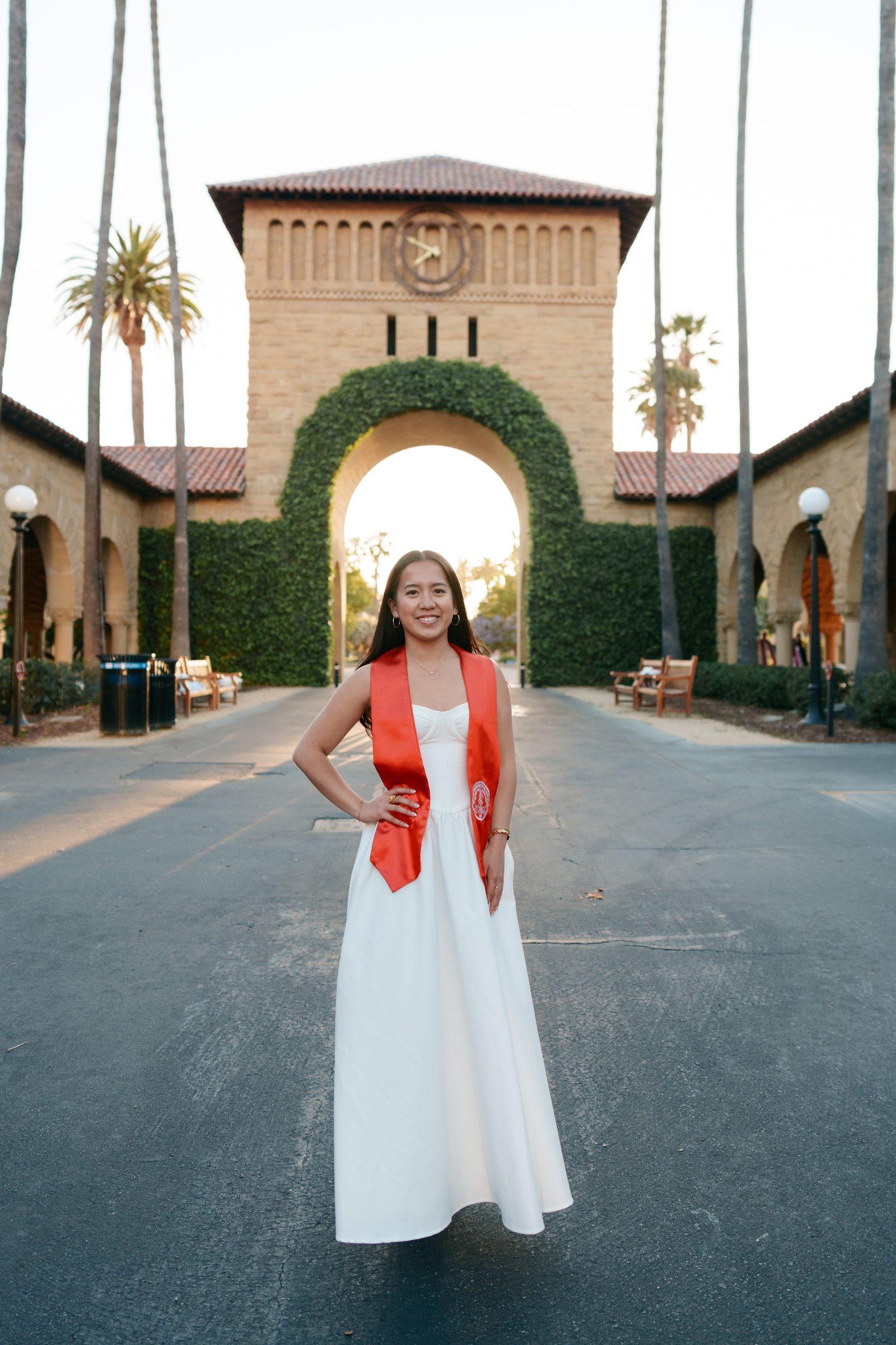 Graduate in white floor-length gown and red stole stands centered in front of an ivy-covered arch and clock tower at golden hour, palm trees framing the shot symmetrically