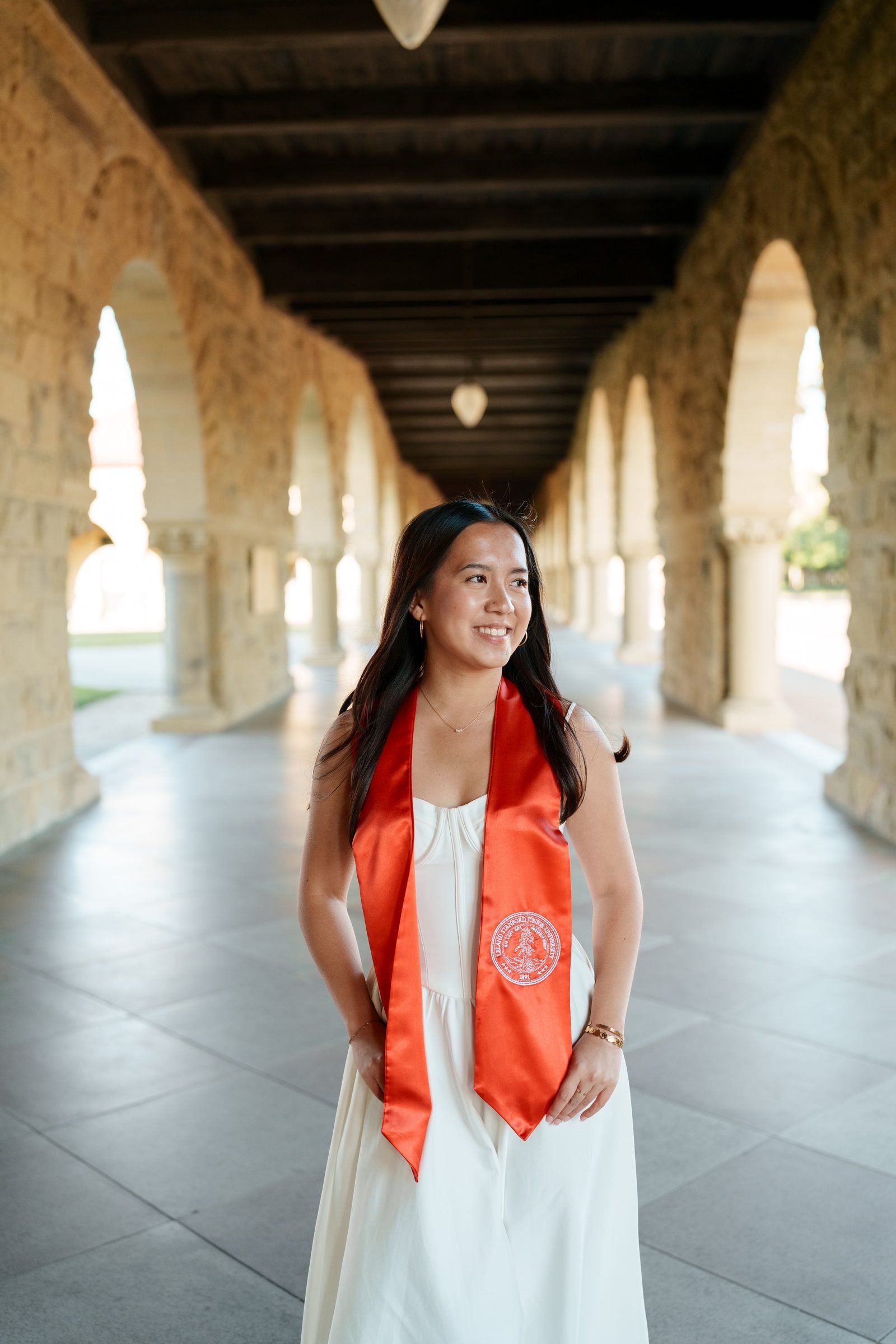 Chantal looks off to the side with a soft smile while standing in Stanford's colonnaded arcade, wearing a white dress and vibrant orange-red graduation stole, warm light filtering through the arches.
