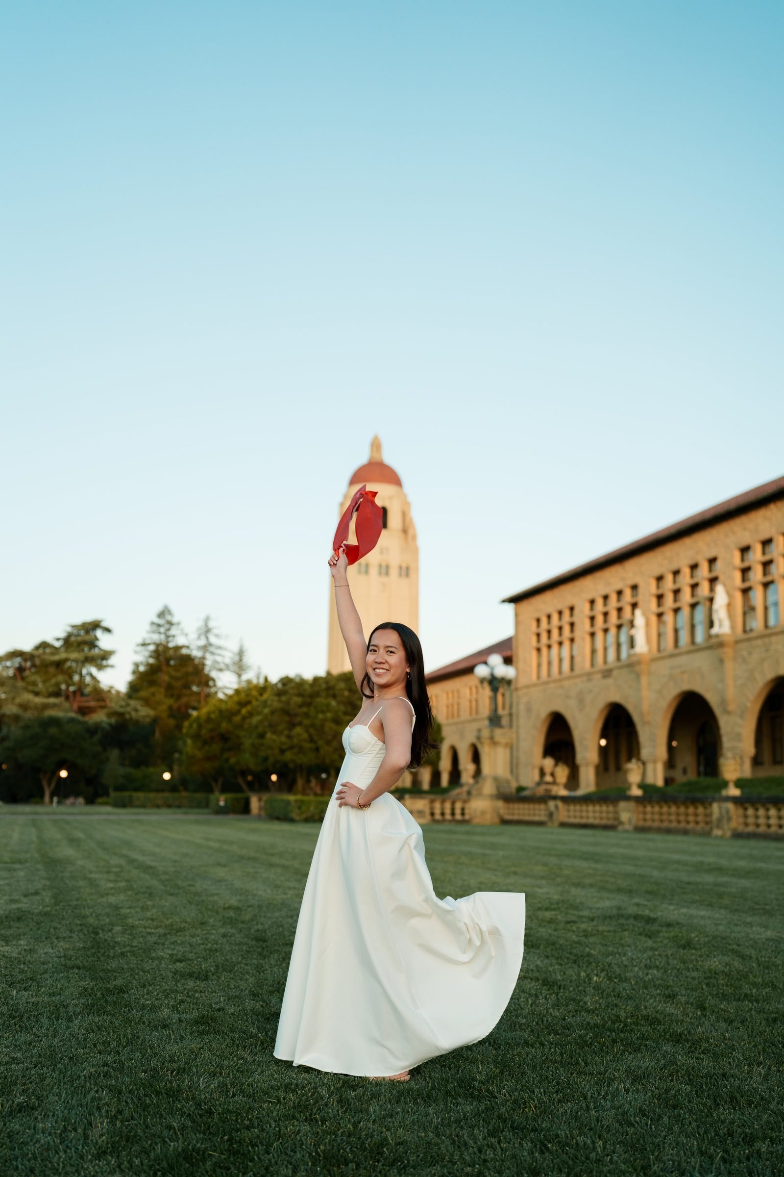 Chantal in a white gown raises her cardinal red graduation stole overhead on the Main Quad lawn at golden hour, Hoover Tower glowing in the soft evening light behind her.