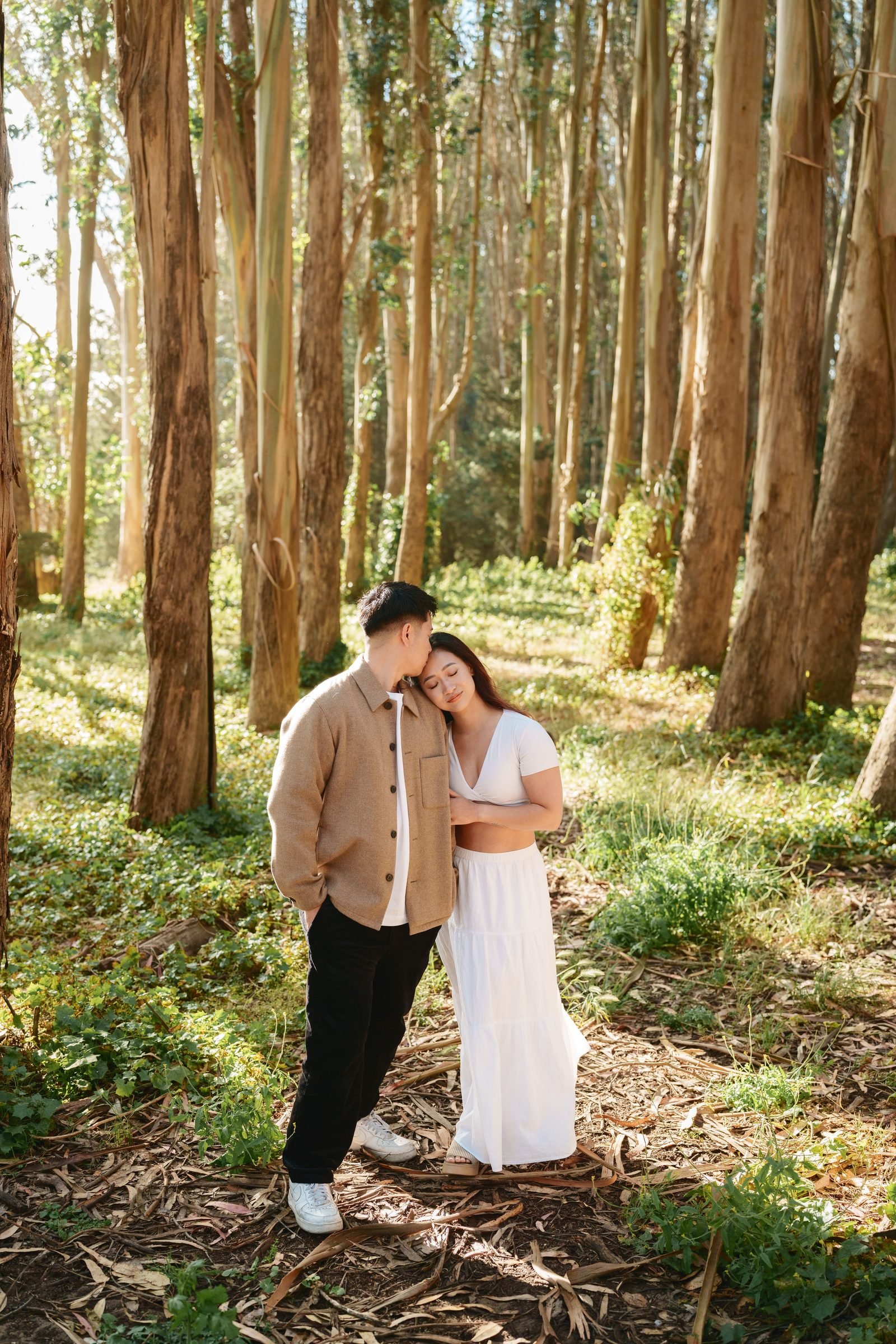 A couple stands still in a eucalyptus grove bathed in soft golden light. She rests her head on his shoulder with eyes closed; he gently kisses her temple. She's in white, he's in a camel shacket.