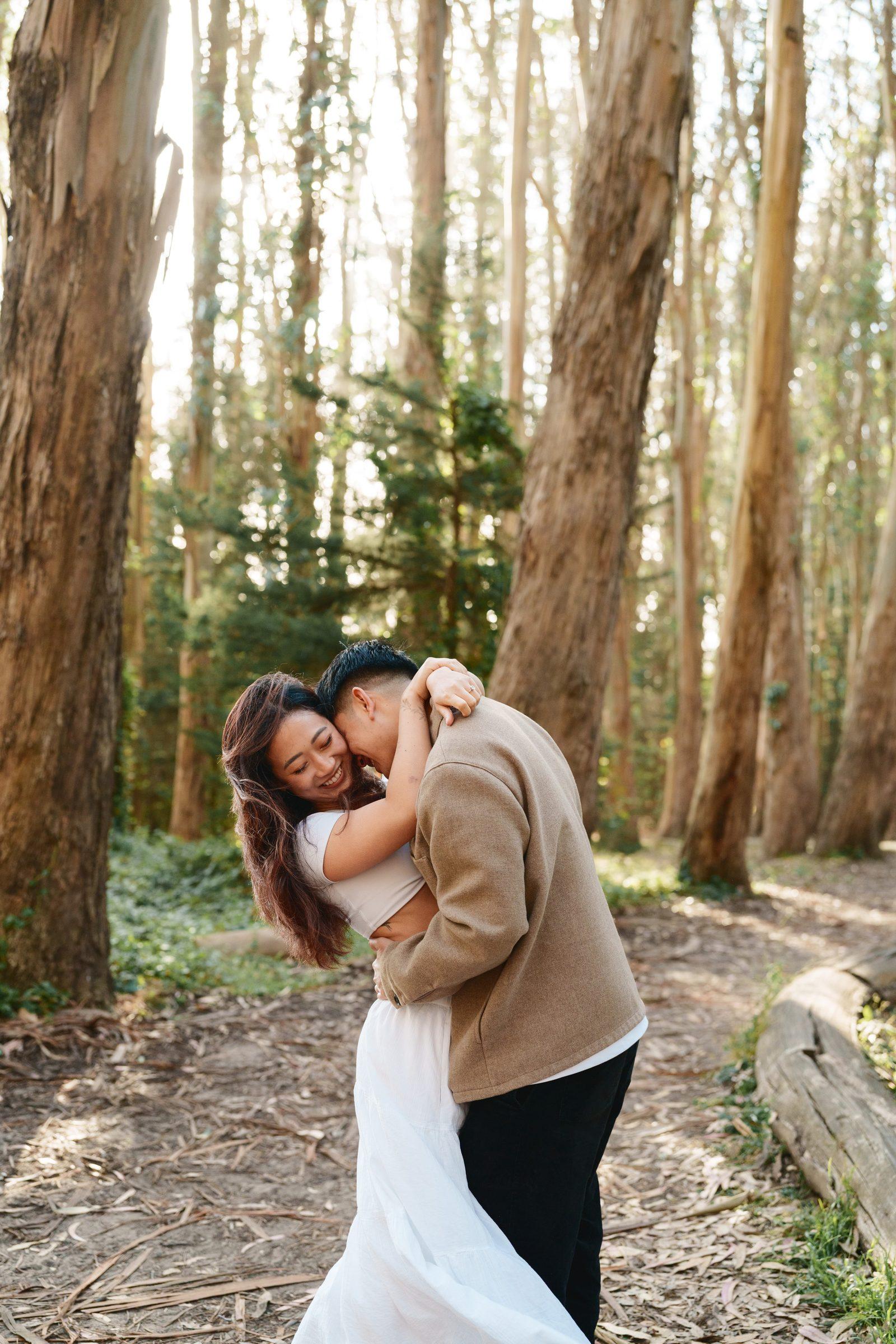 Man dipping and nuzzling his partner in a eucalyptus grove, her head thrown back in a full laugh, white skirt swirling, with tall warm-toned tree trunks and soft dappled light filling the background.