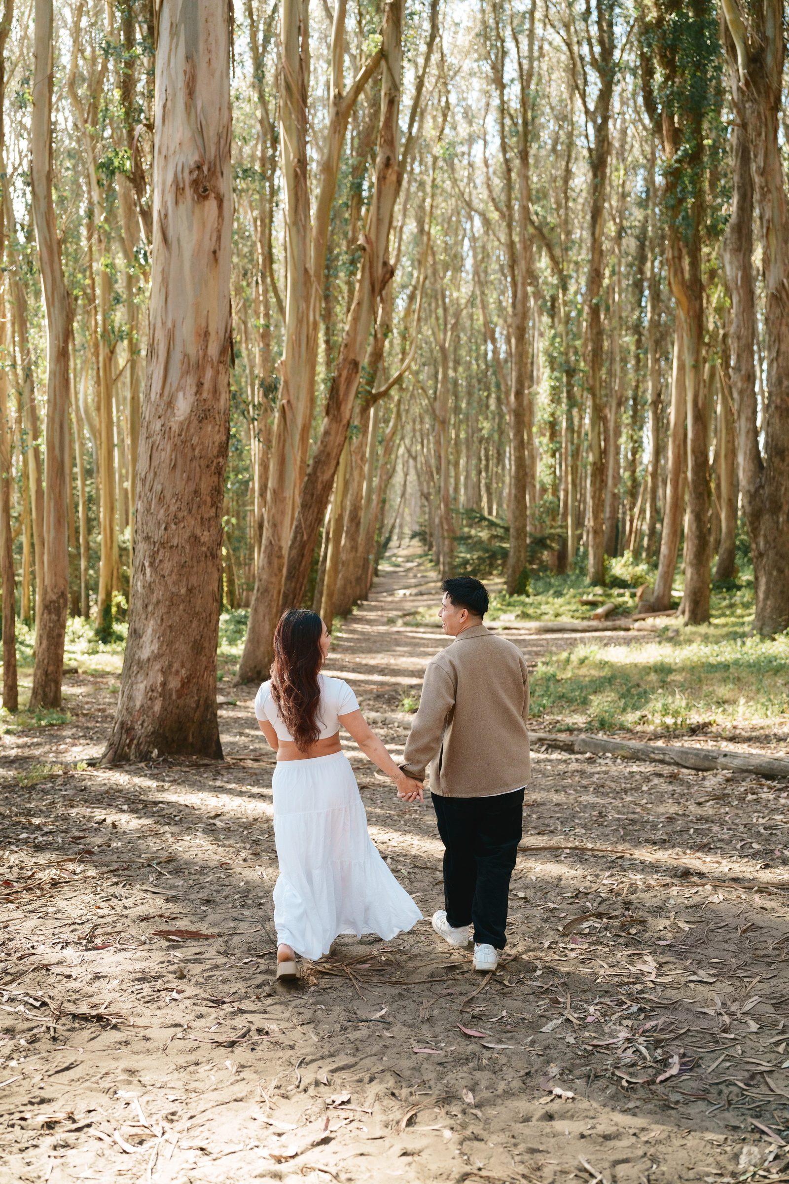 A couple walks away from camera hand in hand into a long eucalyptus grove path, surrounded by tall golden-barked trees and dappled sunlight. She wears a white crop and maxi skirt, he wears a camel jacket.