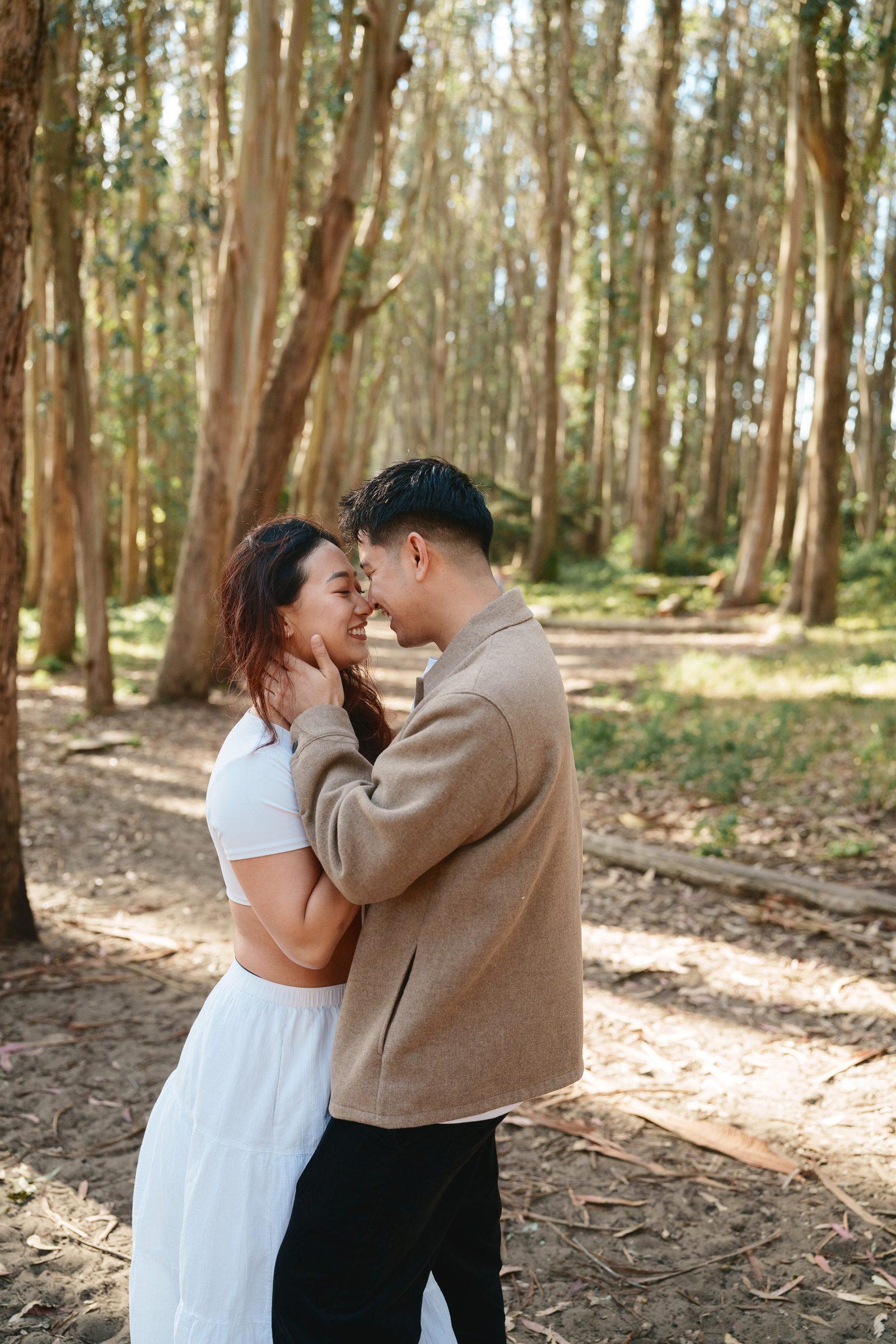 A couple shares a forehead-to-forehead moment on a sun-dappled eucalyptus grove path, her laughing warmly as he cups her face. She wears a white crop top and skirt; he wears a tan jacket.