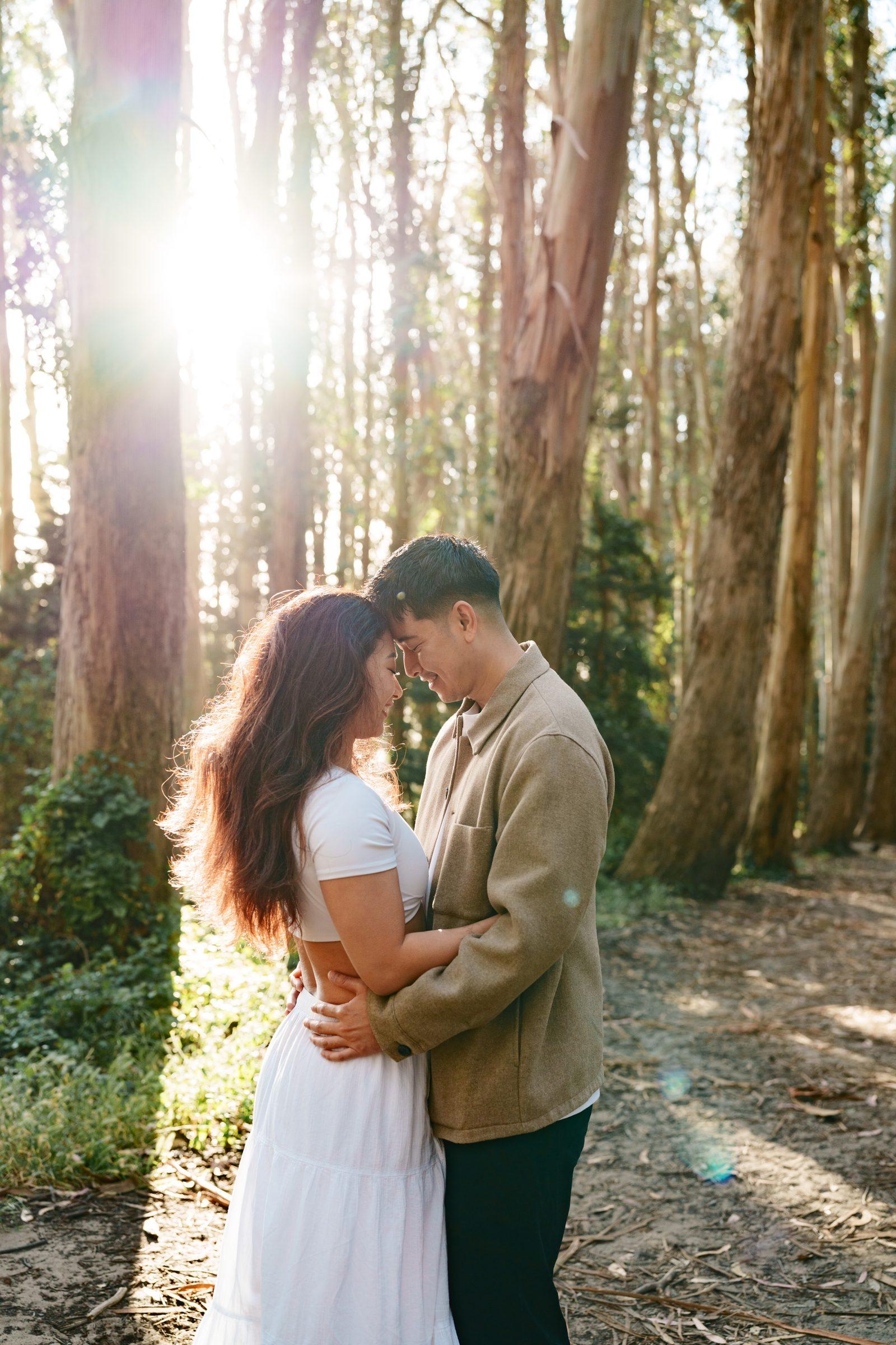 Couple embracing closely in a eucalyptus forest, noses almost touching and both smiling softly, with a dramatic burst of golden backlight and colorful lens flare glowing between the tall tree trunks behind them.
