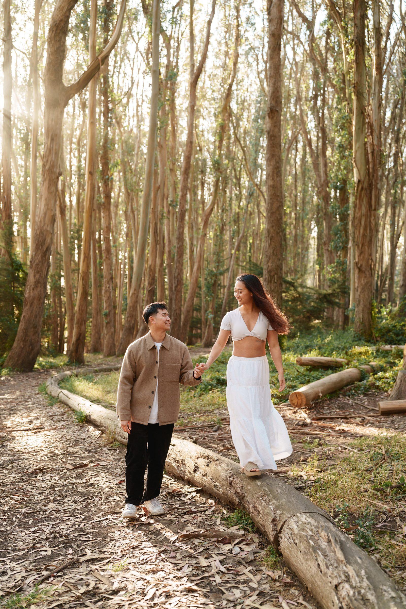 Couple walking hand in hand through a sunlit eucalyptus forest, she balances on a fallen log in a white crop top and flowing skirt, hair catching the breeze, while he looks up at her smiling in a tan button-up jacket and black pants.