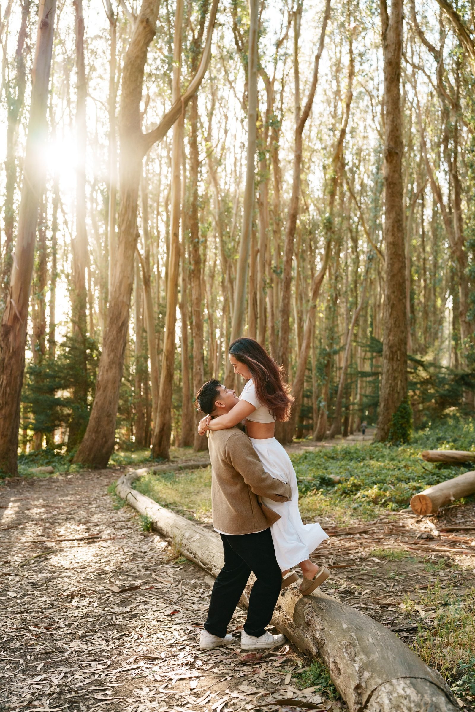 A man lifts a woman off a fallen log, her arms around his neck and her skirt swaying, as bright sun bursts through the eucalyptus canopy directly behind them. The forest floor is scattered with dried bark and green undergrowth.