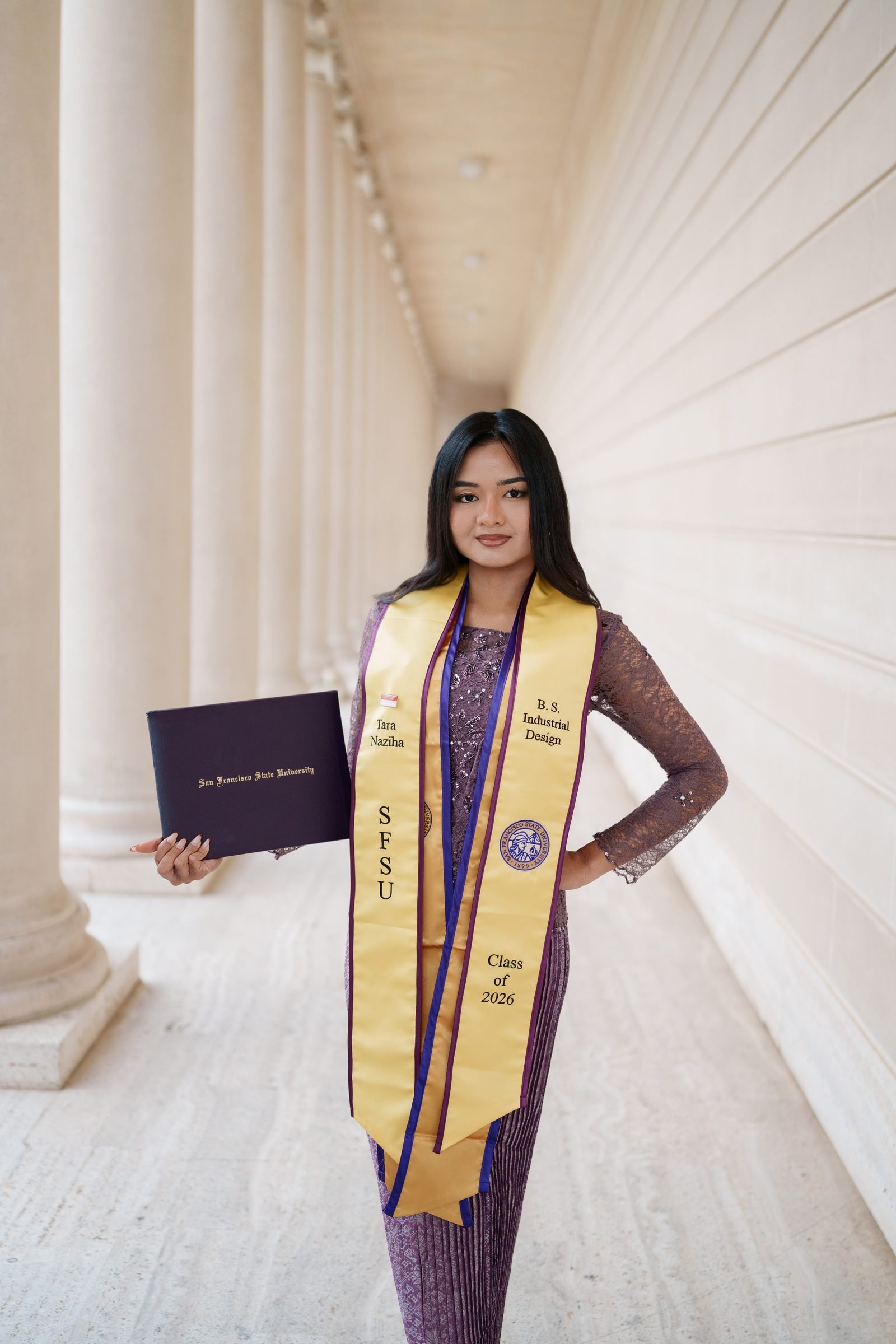 Tara poses with one hand on her hip holding her SFSU diploma cover in the Legion of Honor colonnade, wearing a purple lace dress and gold graduation stole, bathed in even midday light.