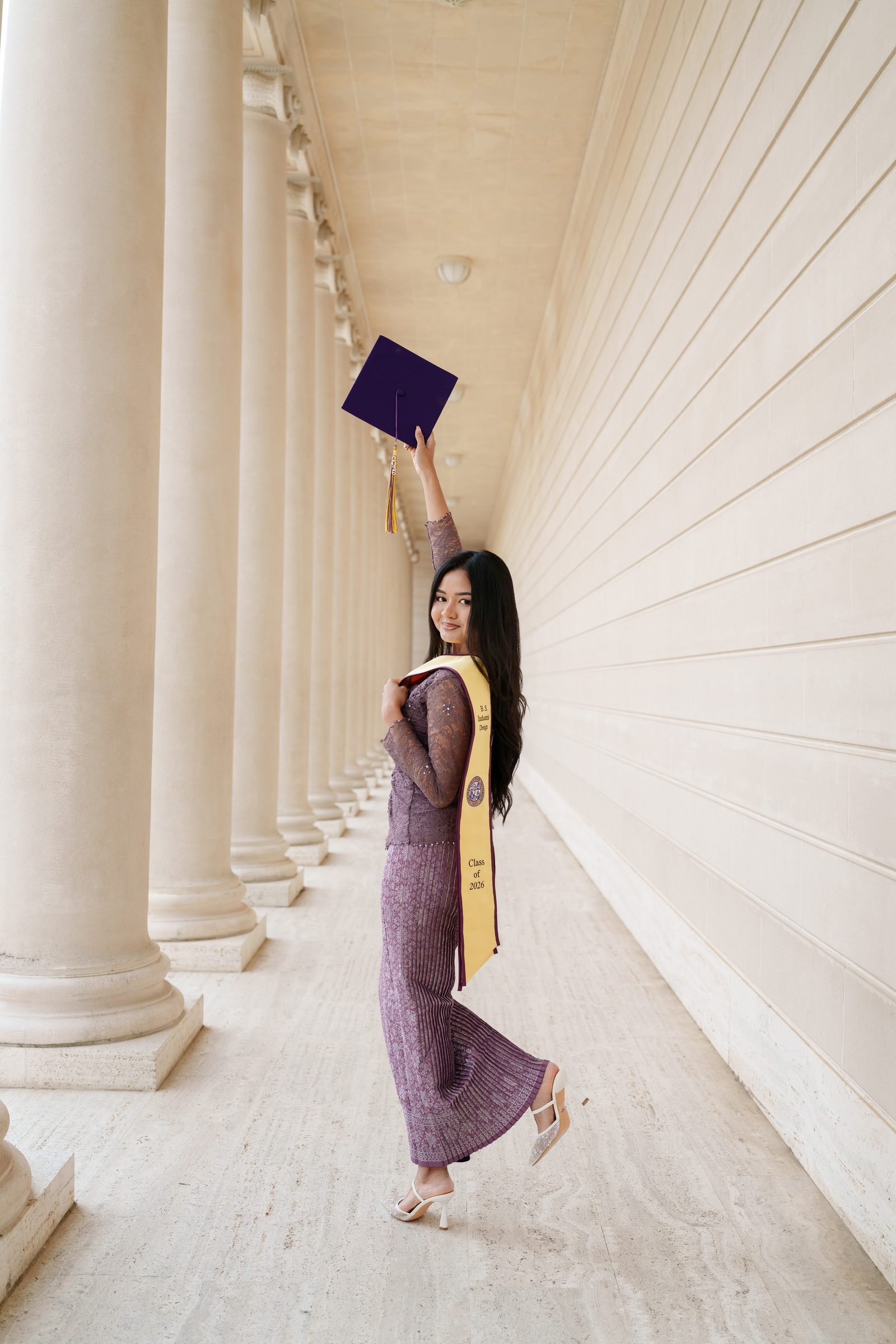 Tara holds her graduation cap high above her head while glancing back over her shoulder beneath the long colonnade at the Legion of Honor, wearing a purple lace outfit and gold Class of 2026 stole.