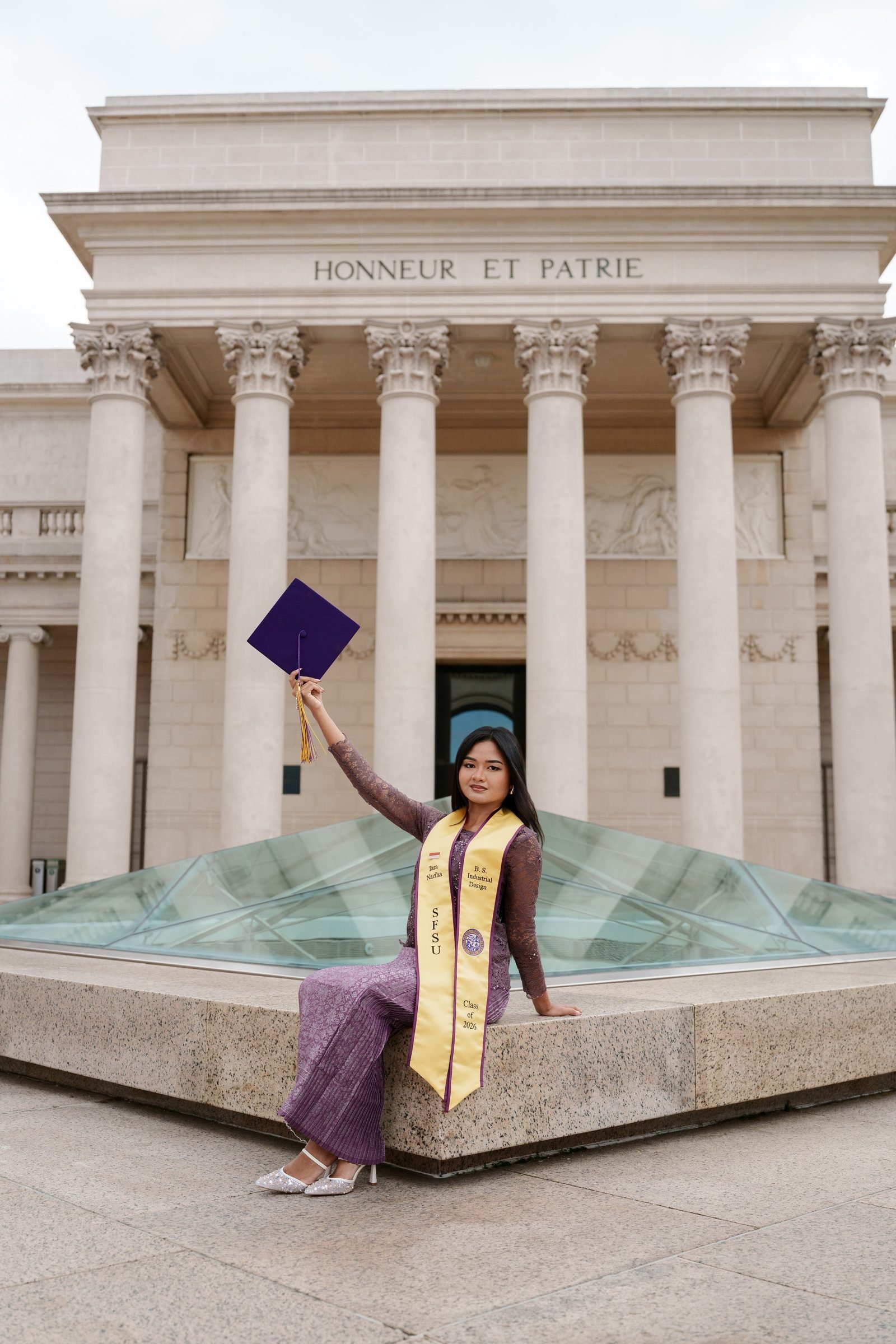 Tara sits on the stone ledge outside the Legion of Honor and raises her purple mortarboard high overhead with one arm, smiling, with the grand columned building filling the background under an overcast sky.