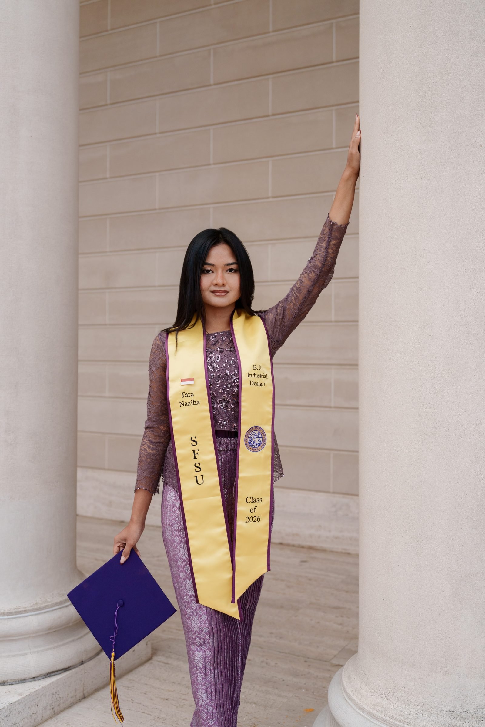 Tara stands between two tall columns on the Legion of Honor portico, one hand raised against the stone pillar, holding her purple mortarboard at her side, wearing a purple lace kebaya and gold SFSU stole in soft diffused light.