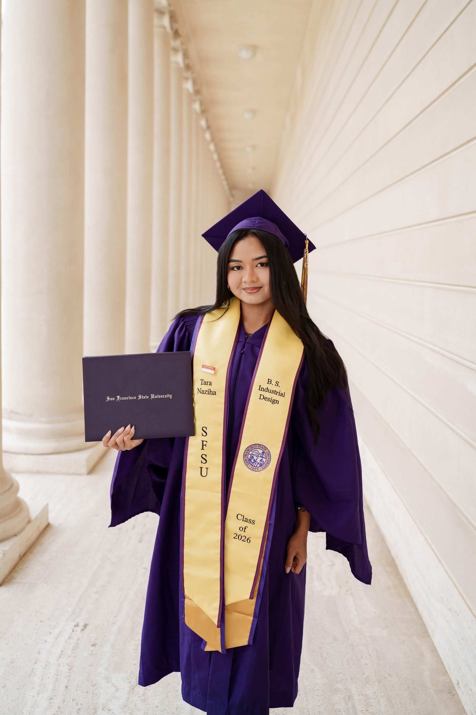 SFSU graduate Tara stands in the colonnaded corridor of the Legion of Honor, holding her diploma cover in her purple cap and gown with gold stole, warm midday light reflecting off the pale stone columns behind her.