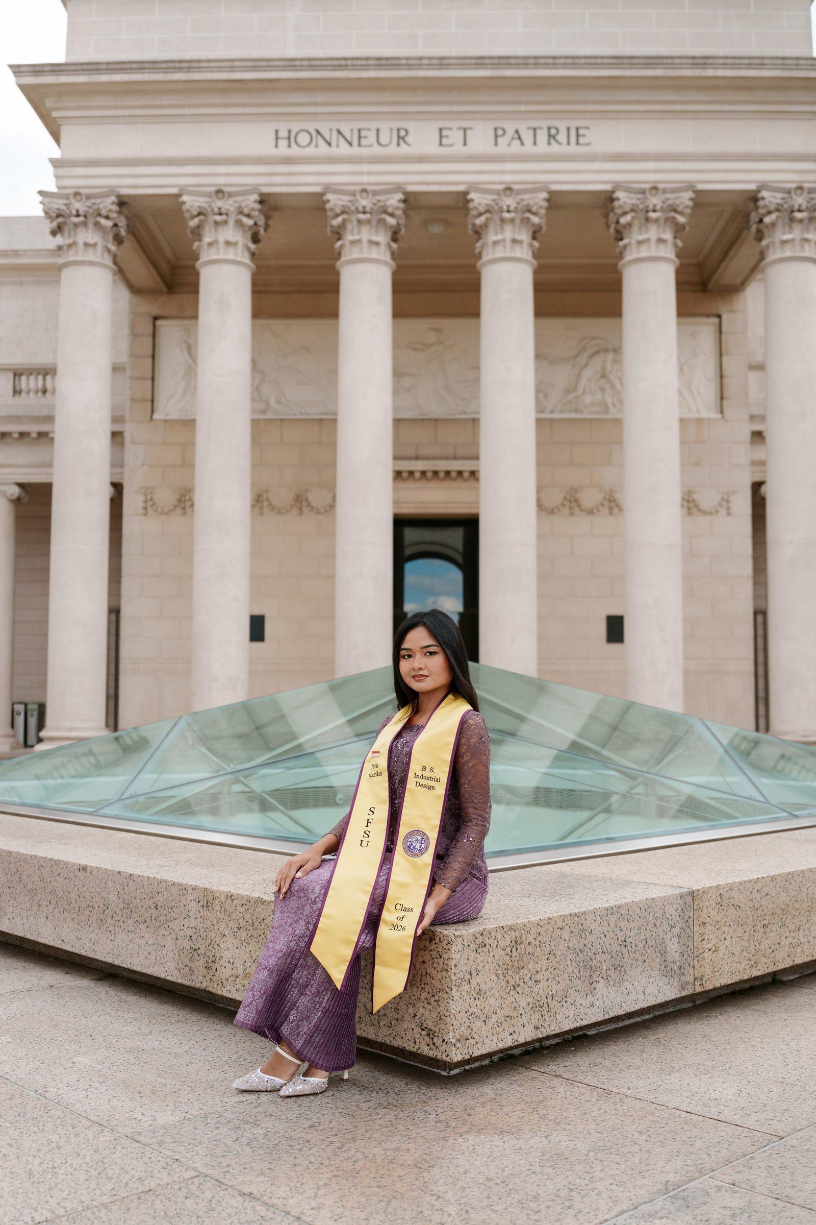 Tara sits in front of the Legion of Honor's columned facade with 'Honneur et Patrie' engraved above her. She wears a purple lace outfit and gold SFSU graduation sash, the glass pyramid skylight visible behind her.