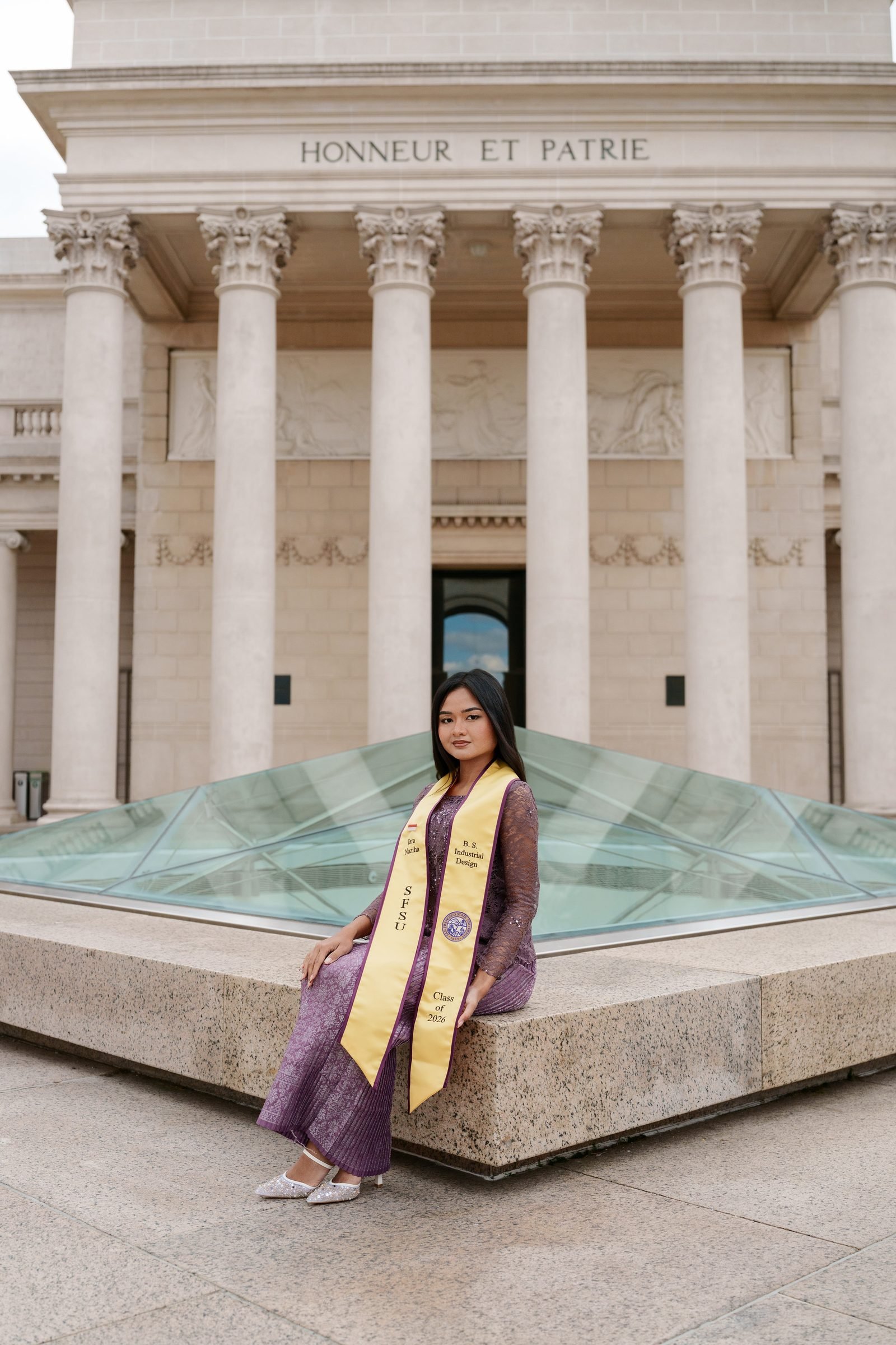 Tara sits in front of the Legion of Honor's columned facade with 'Honneur et Patrie' engraved above her. She wears a purple lace outfit and gold SFSU graduation sash, the glass pyramid skylight visible behind her.