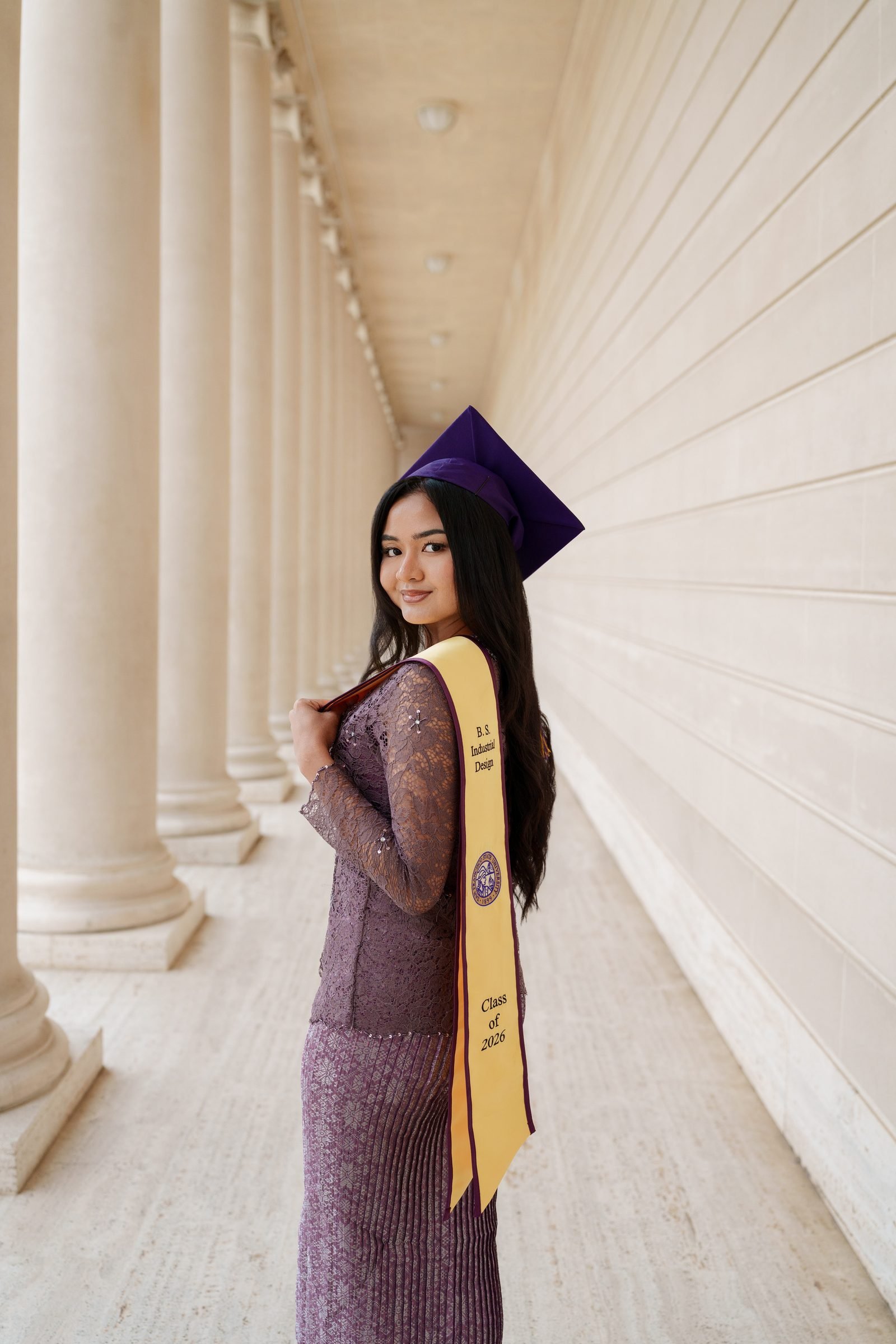 Tara looks back over her shoulder at the camera wearing her purple mortarboard and gold B.S. Industrial Design stole, standing in the covered colonnade at the Legion of Honor with soft diffused light wrapping around her.