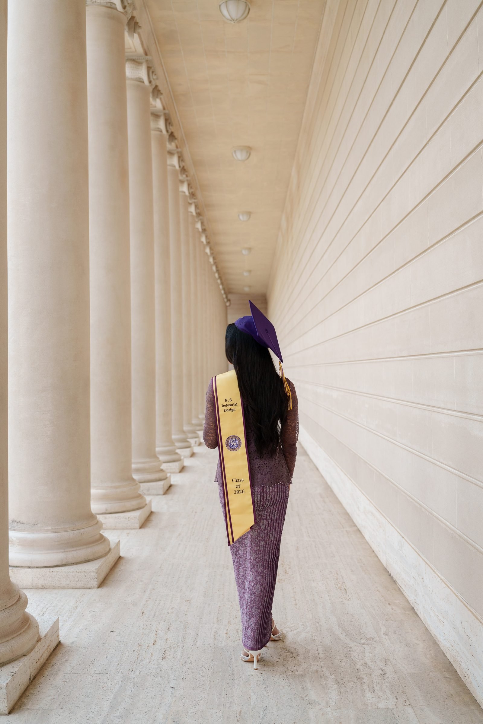 Tara walks away from the camera down the Legion of Honor colonnade, her back to the lens, purple cap and gold B.S. Industrial Design stole visible as the columns and cream walls recede into the distance.