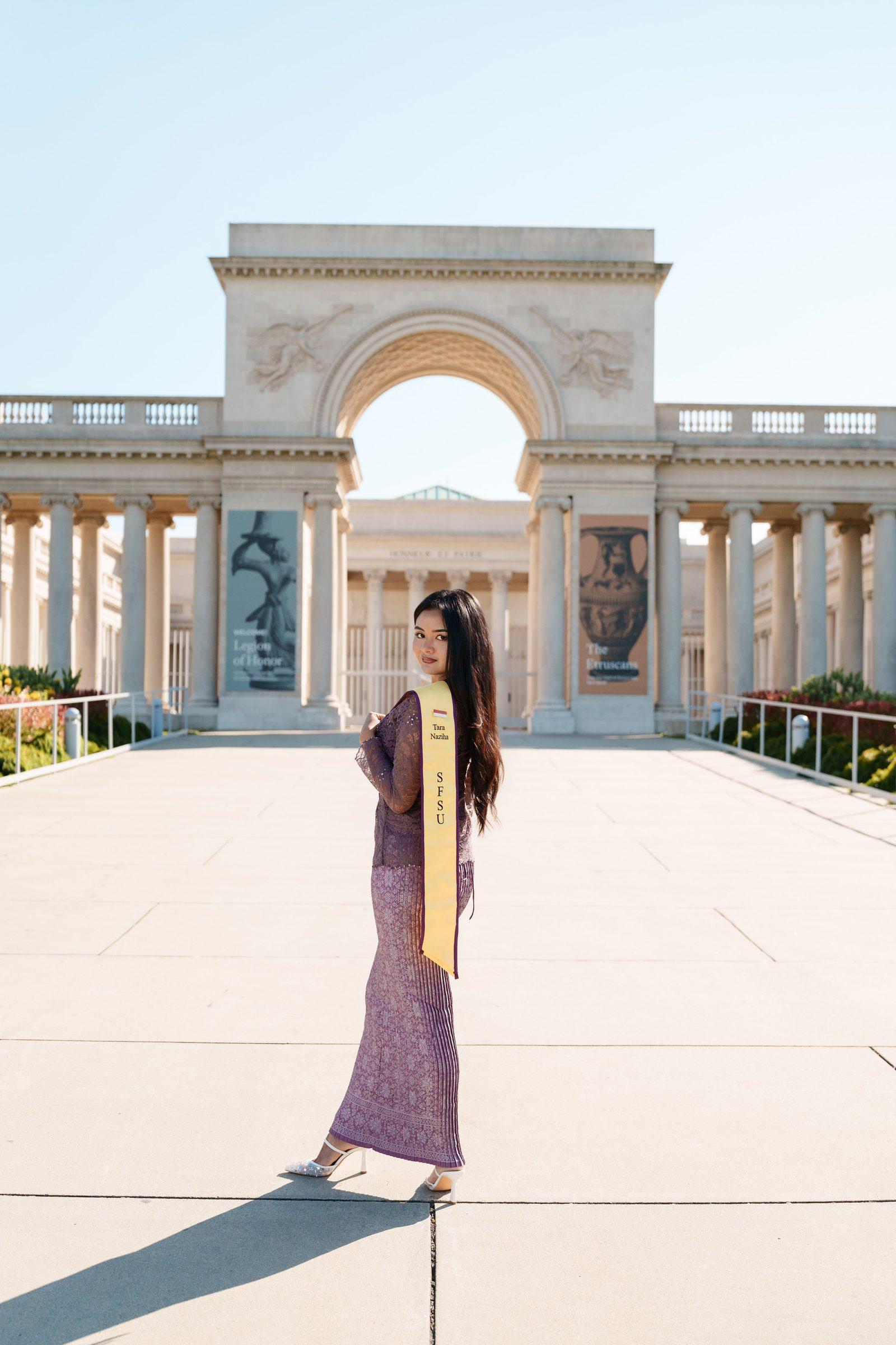 Tara stands on the wide stone promenade in front of the Legion of Honor's neoclassical arch, wearing a floor-length purple lace gown and gold SFSU stash, glancing back over her shoulder in bright midday light.