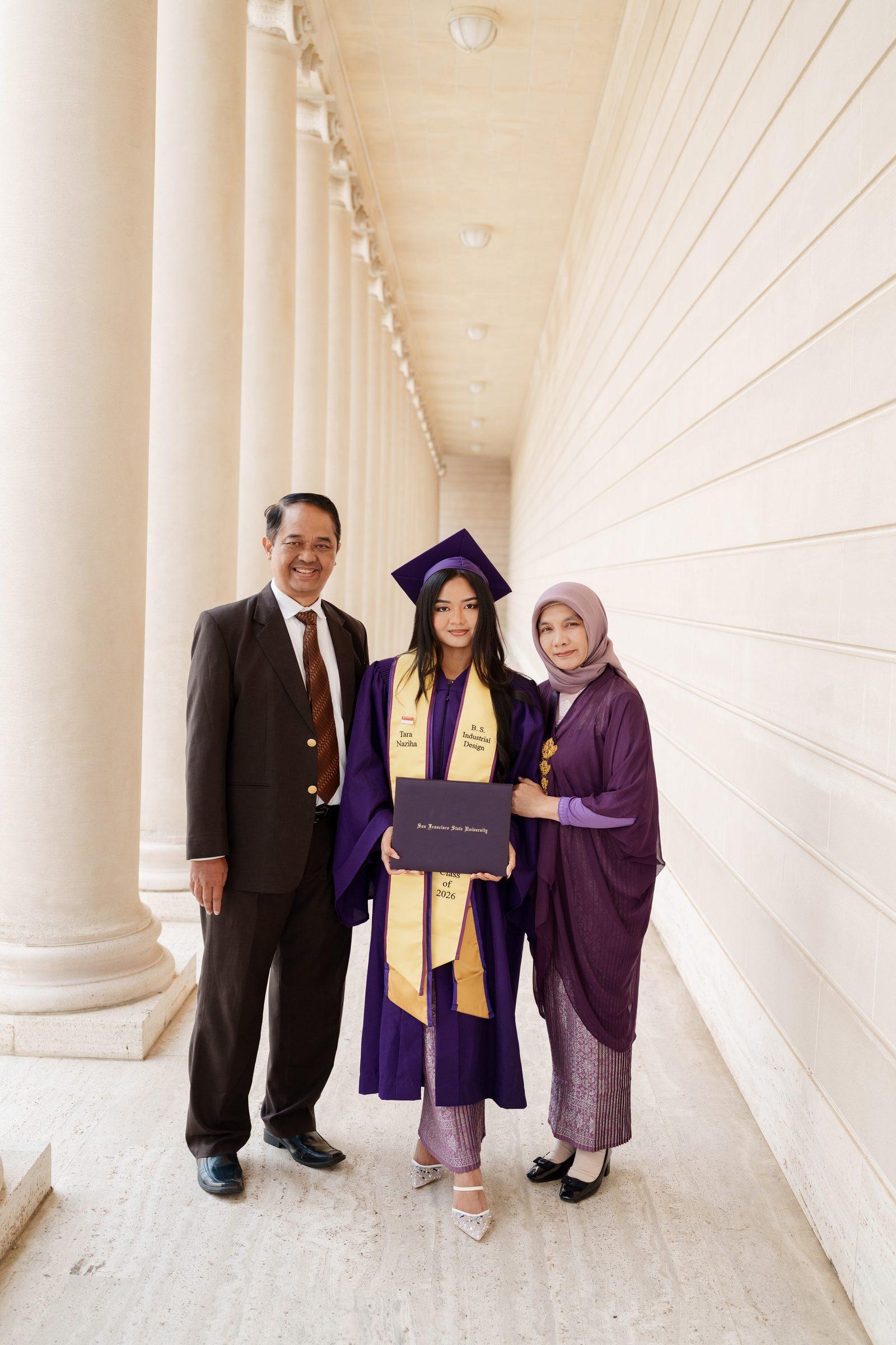 Tara stands between her parents in the Legion of Honor colonnade — her father in a dark suit, her mother in a purple hijab and traditional kebaya — all three smiling warmly as Tara holds her SFSU diploma.