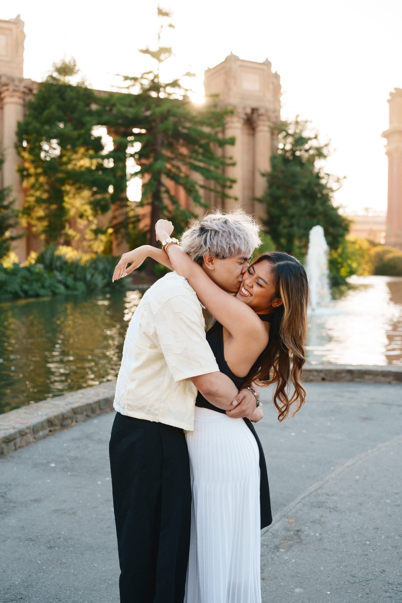A couple embraces and laughs at the Palace of Fine Arts during golden hour, the lagoon and fountain glowing behind them. One wears a cream shirt with black trousers, the other a black top with a white pleated skirt.