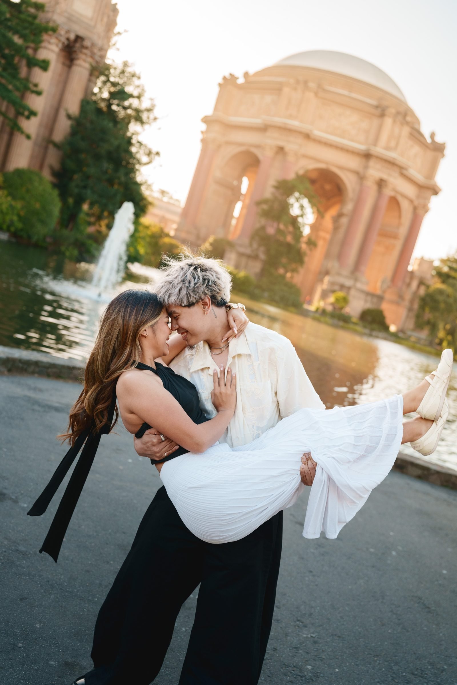 One partner carries the other in their arms as they lean foreheads together and smile at Palace of Fine Arts during golden hour, warm light glowing on the rotunda behind them.