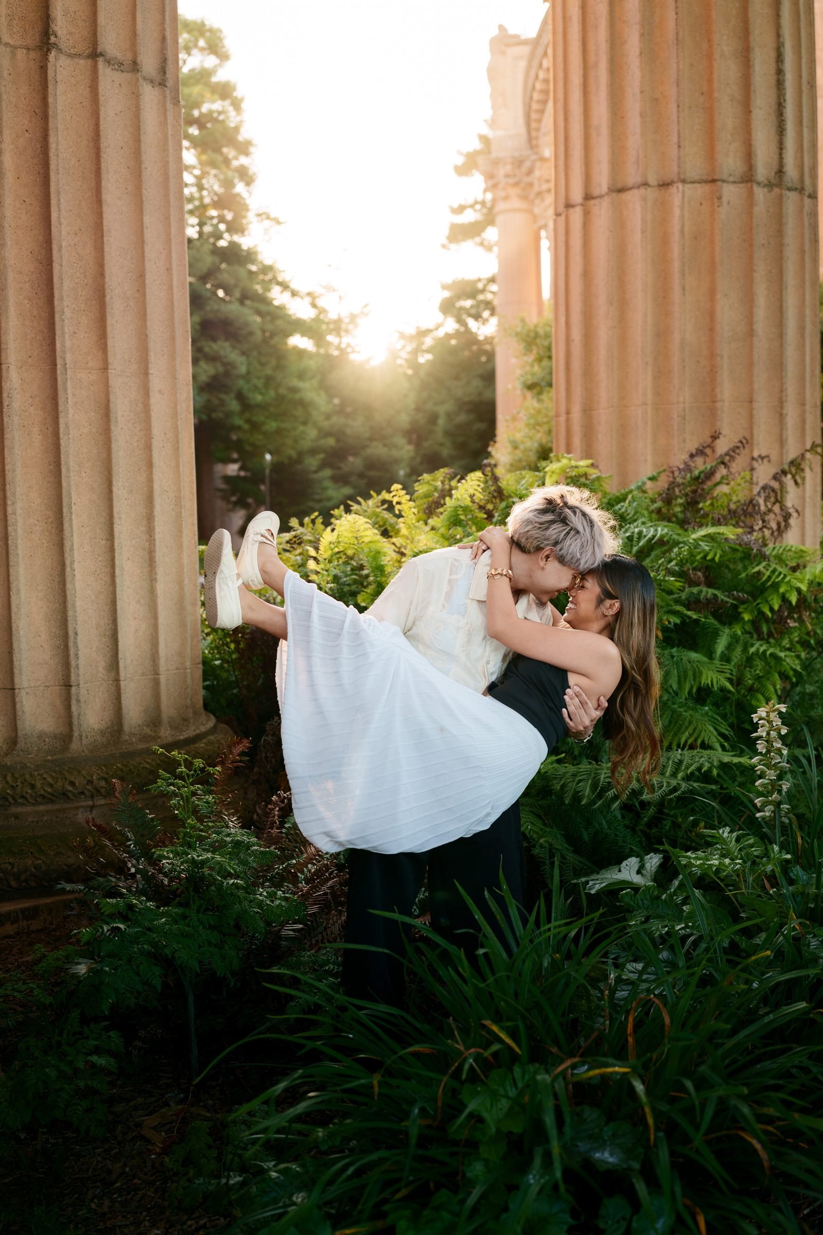 A couple shares a joyful kiss as one partner lifts the other among lush ferns between towering sandstone columns at the Palace of Fine Arts, bathed in golden hour backlight.