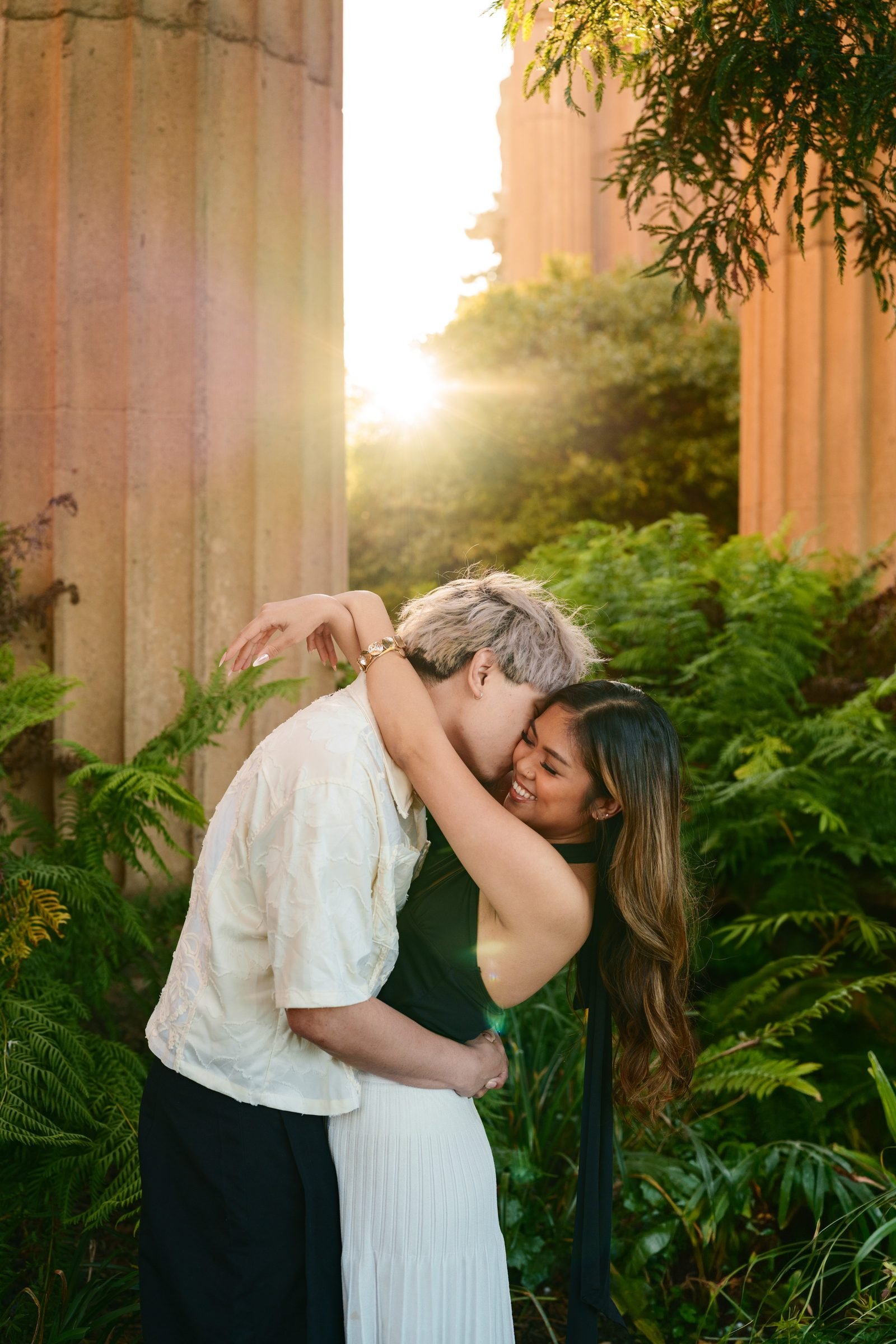A couple embraces between sandstone columns at the Palace of Fine Arts during golden hour, surrounded by lush ferns. He nuzzles into her neck while she laughs, wearing a green top and white skirt; he wears a cream patterned shirt.