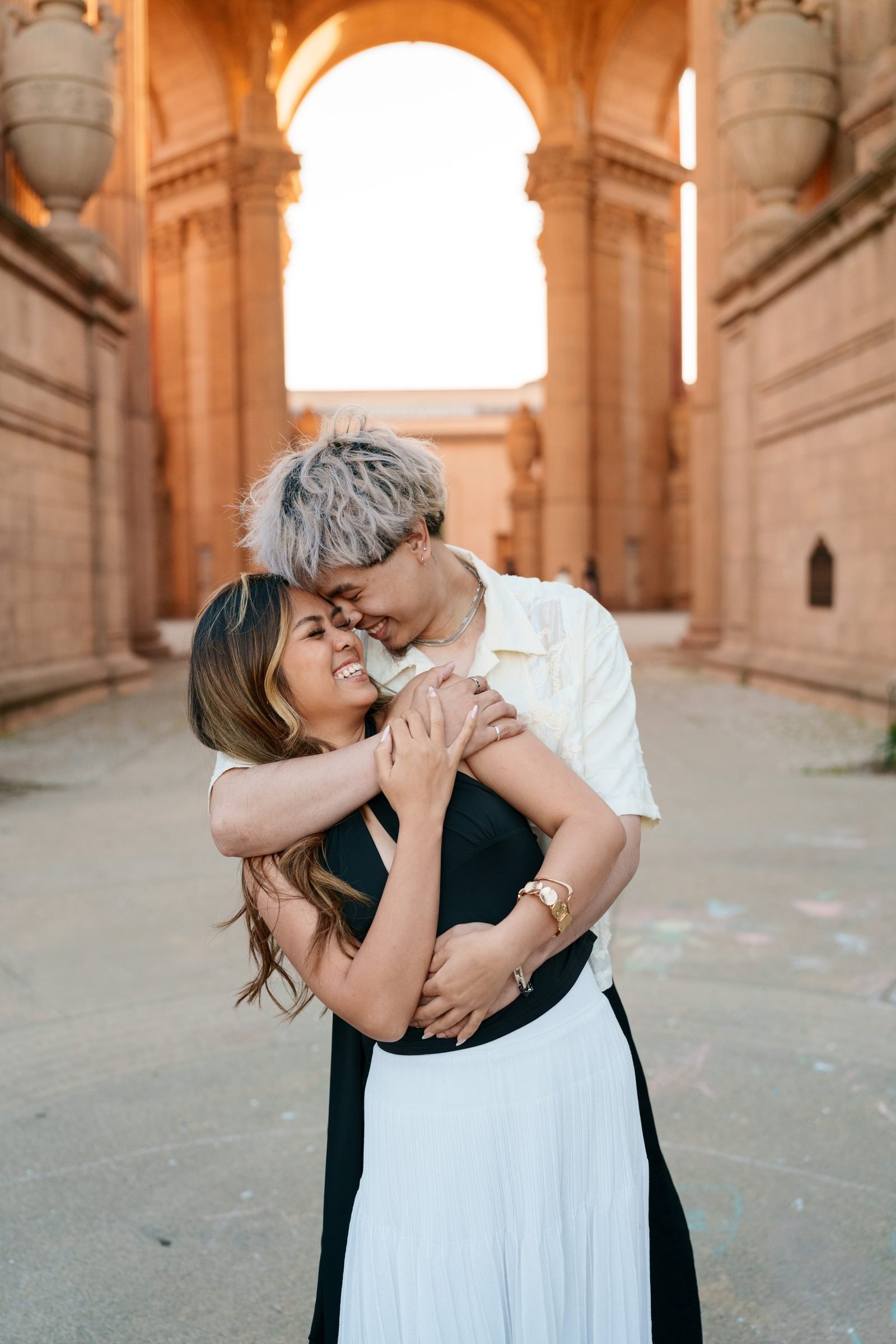 A couple embraces warmly inside the colonnade of the Palace of Fine Arts during golden hour, the warm terracotta arches glowing behind them as she laughs and he nuzzles close, both in coordinated black, white, and cream tones.