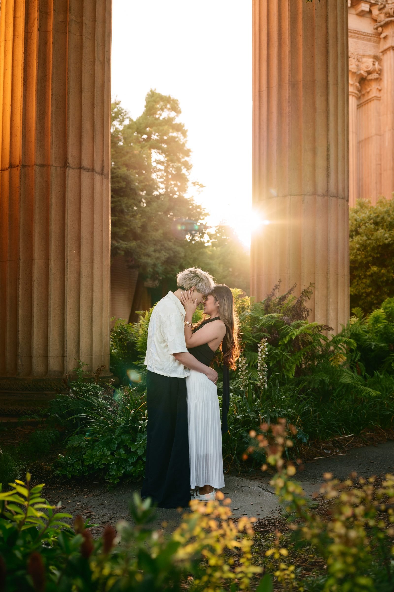 A couple stands forehead-to-forehead between massive sandstone columns at the Palace of Fine Arts, bathed in golden hour backlight with a starburst sun and lush greenery surrounding them.