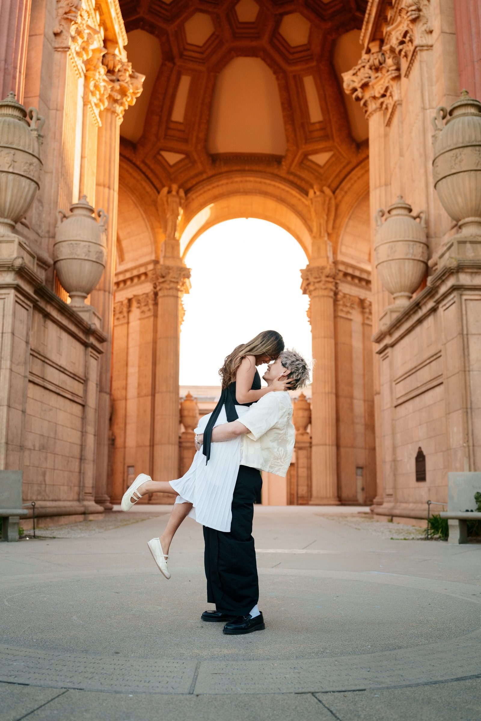 A couple share a lifted kiss beneath the grand arched colonnade of the Palace of Fine Arts during golden hour, bathed in warm terracotta light. One partner wears cream linen and black trousers, the other a white pleated dress with a black ribbon tie.