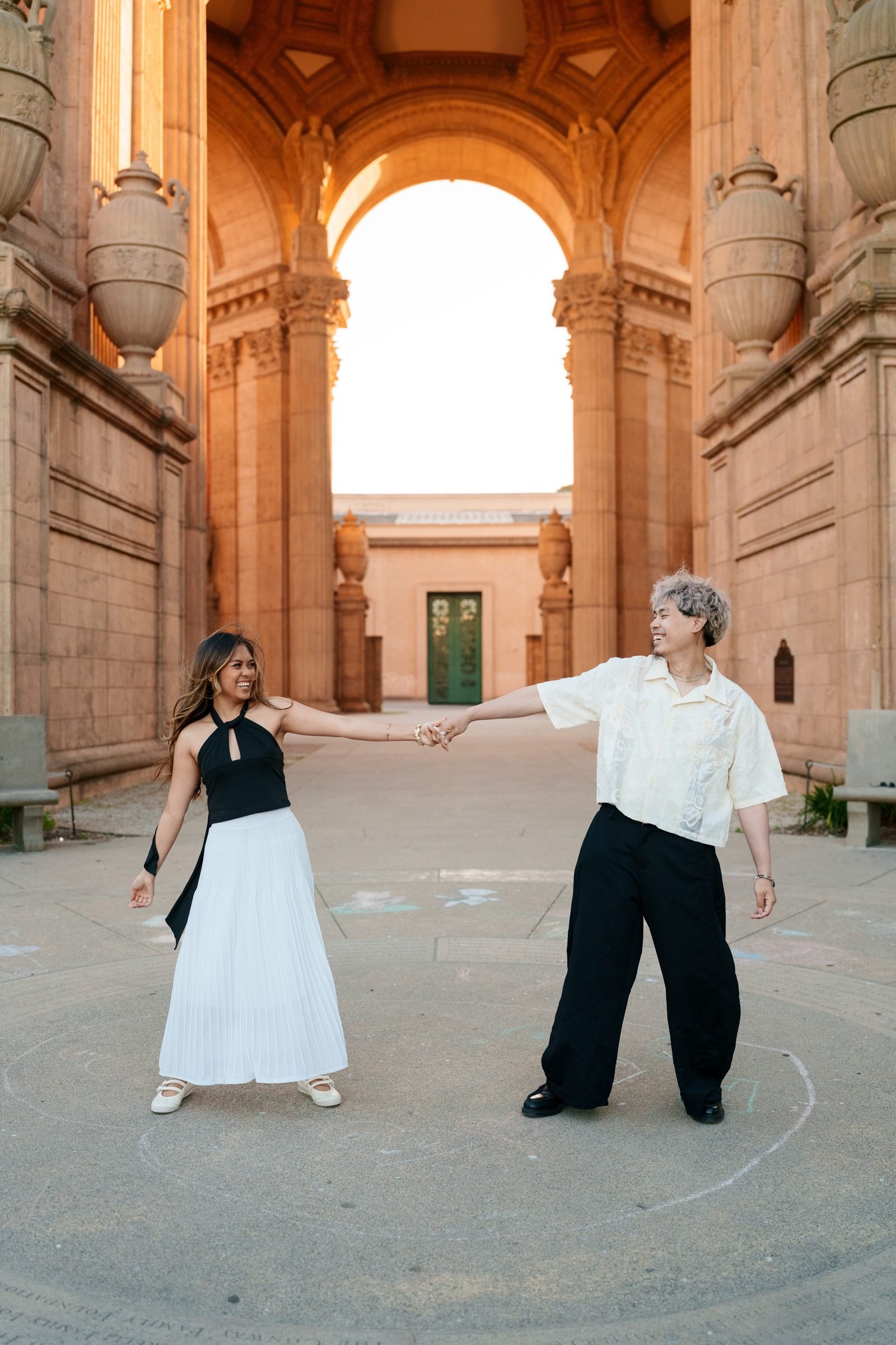 A couple holds hands at arm's length beneath the grand arched colonnade of the Palace of Fine Arts during golden hour, both laughing as warm amber light glows across the terracotta stonework behind them.