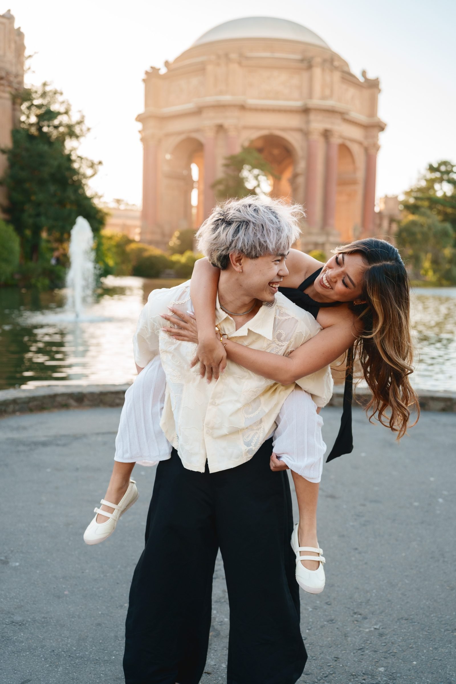 A couple laughs together at the Palace of Fine Arts during golden hour — one giving the other a piggyback ride in front of the domed rotunda and lagoon fountain, wearing cream, white, and black tones.