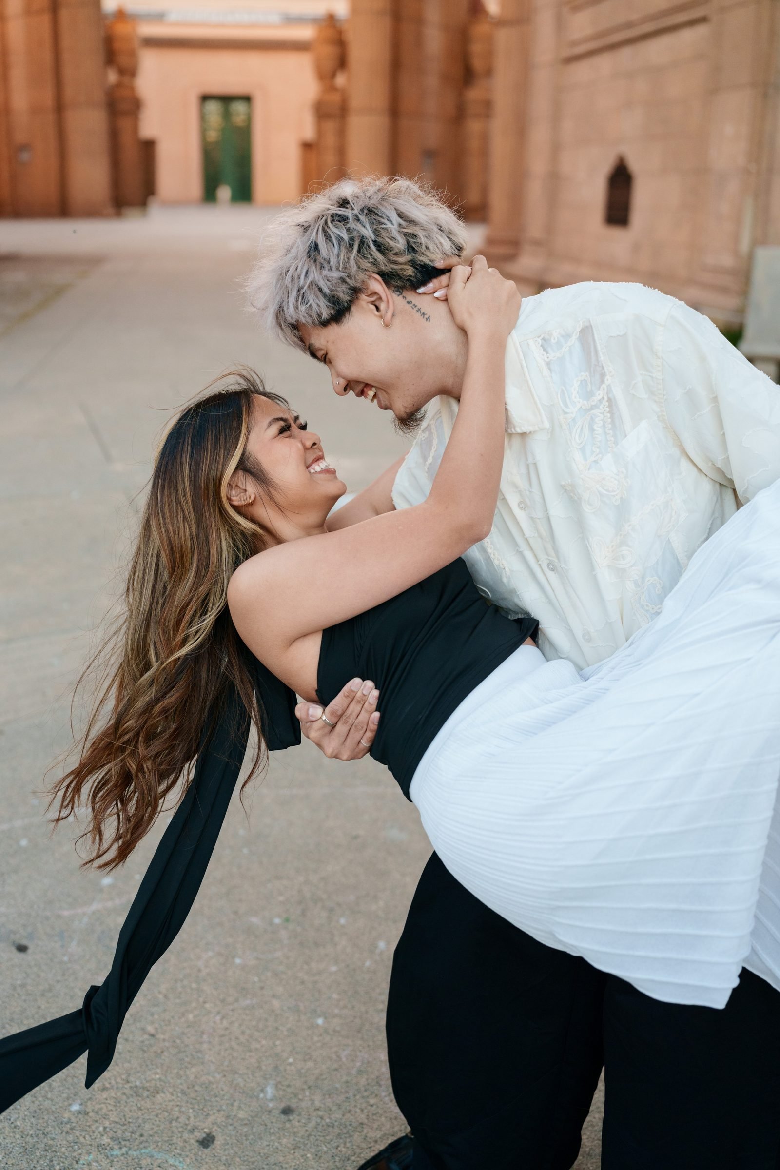 A couple shares a playful dip in front of the warm sandstone columns of the Palace of Fine Arts during golden hour. She laughs up at him, hair sweeping wide, as he leans in close. She wears a black tie-back dress; he wears a cream embroidered shirt.