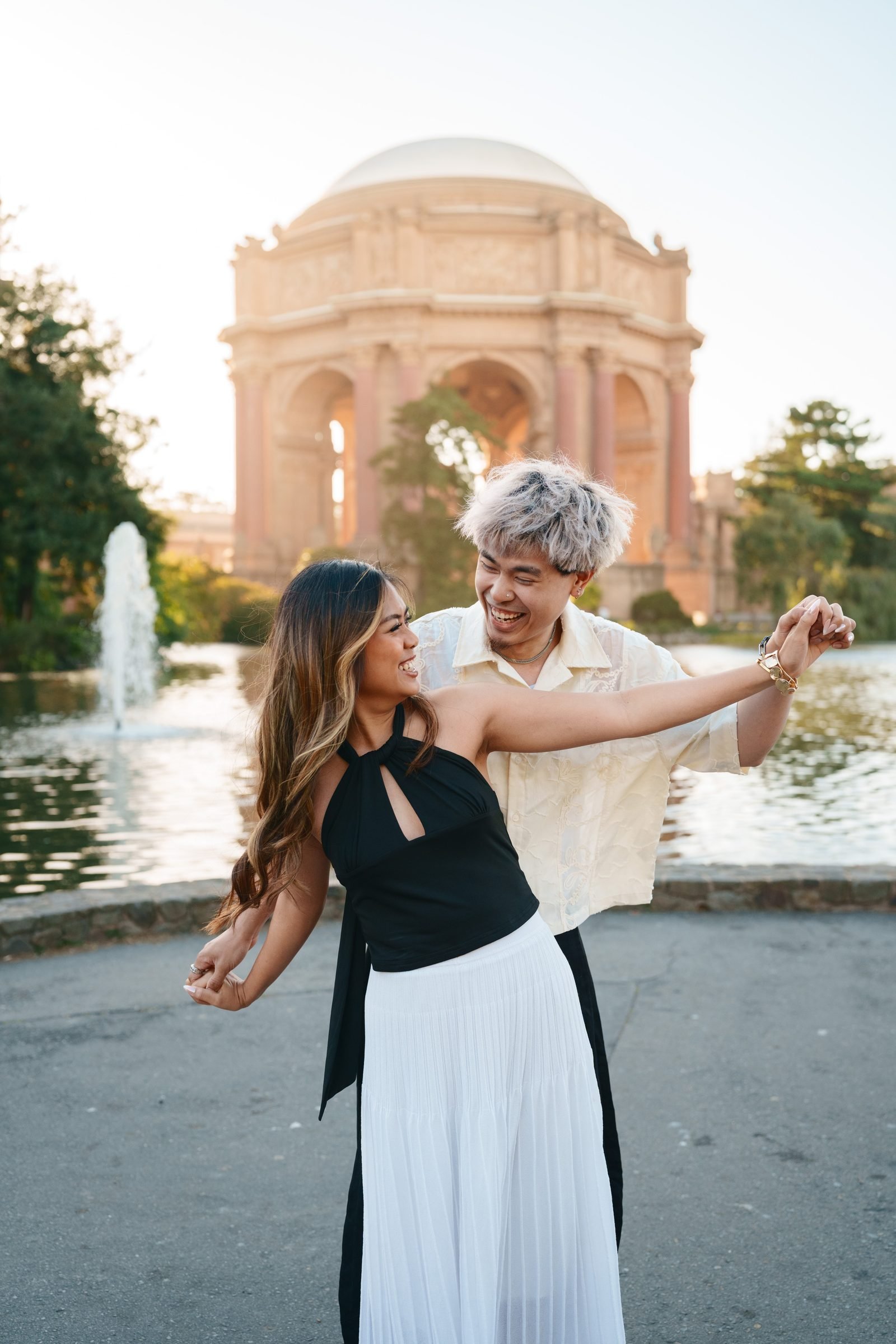 A couple dances and laughs together in front of the Palace of Fine Arts rotunda and reflecting pool during golden hour. She wears a black halter top and white pleated skirt; he wears a cream textured shirt, silver-toned hair.