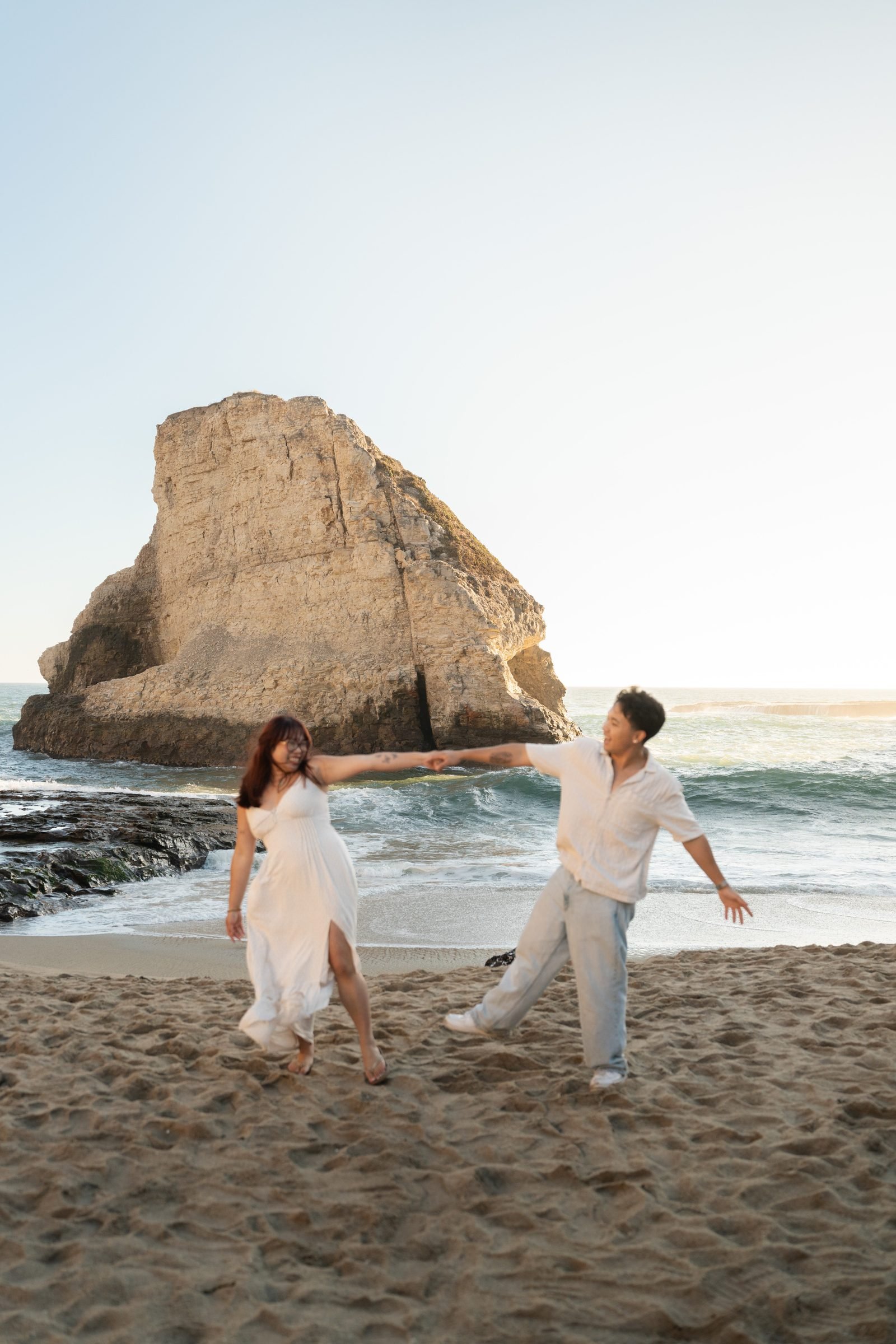 A couple dances playfully on the sandy beach at Shark Fin Cove during golden hour, her white maxi dress catching the breeze as he reaches out to spin her, the iconic sea stack glowing warm behind them.