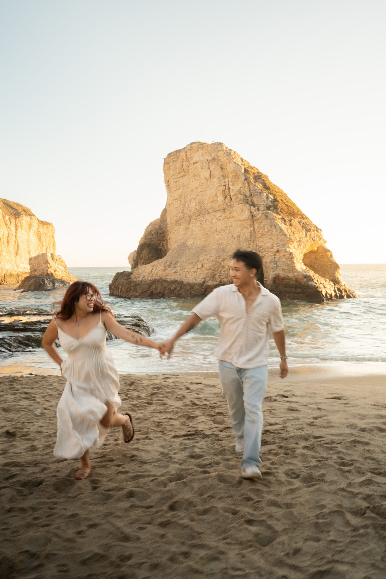 A couple runs hand-in-hand across the sand at Shark Fin Cove during golden hour, laughing freely, she in a white slip dress and he in a white linen shirt and light jeans, the iconic sea stack glowing warm behind them.