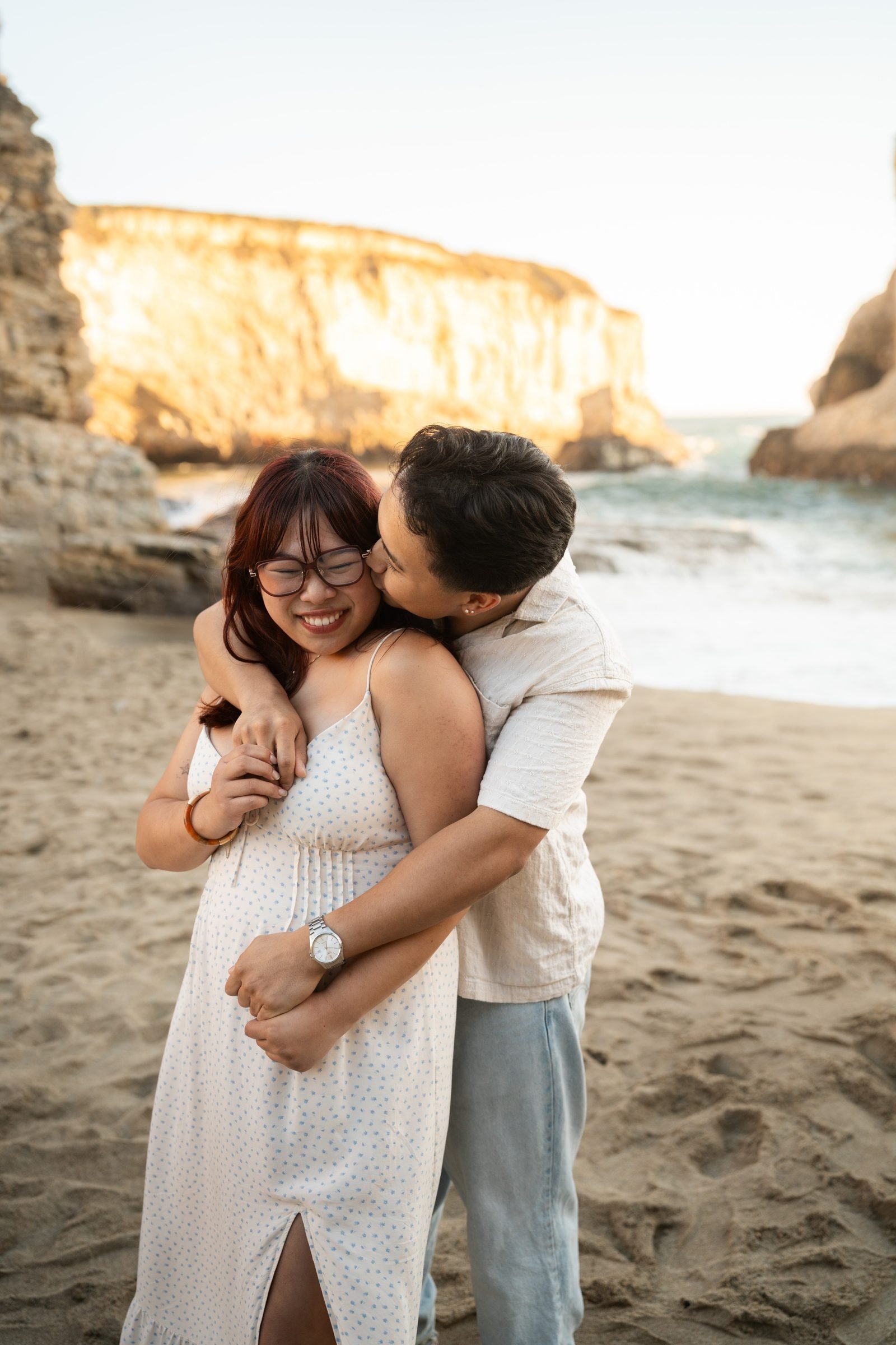 A man wraps his arms around his girlfriend from behind and kisses her cheek as she laughs at Shark Fin Cove during golden hour, warm cliffs glowing in the background, she wears a white floral sundress, he wears a cream linen shirt and light blue pants