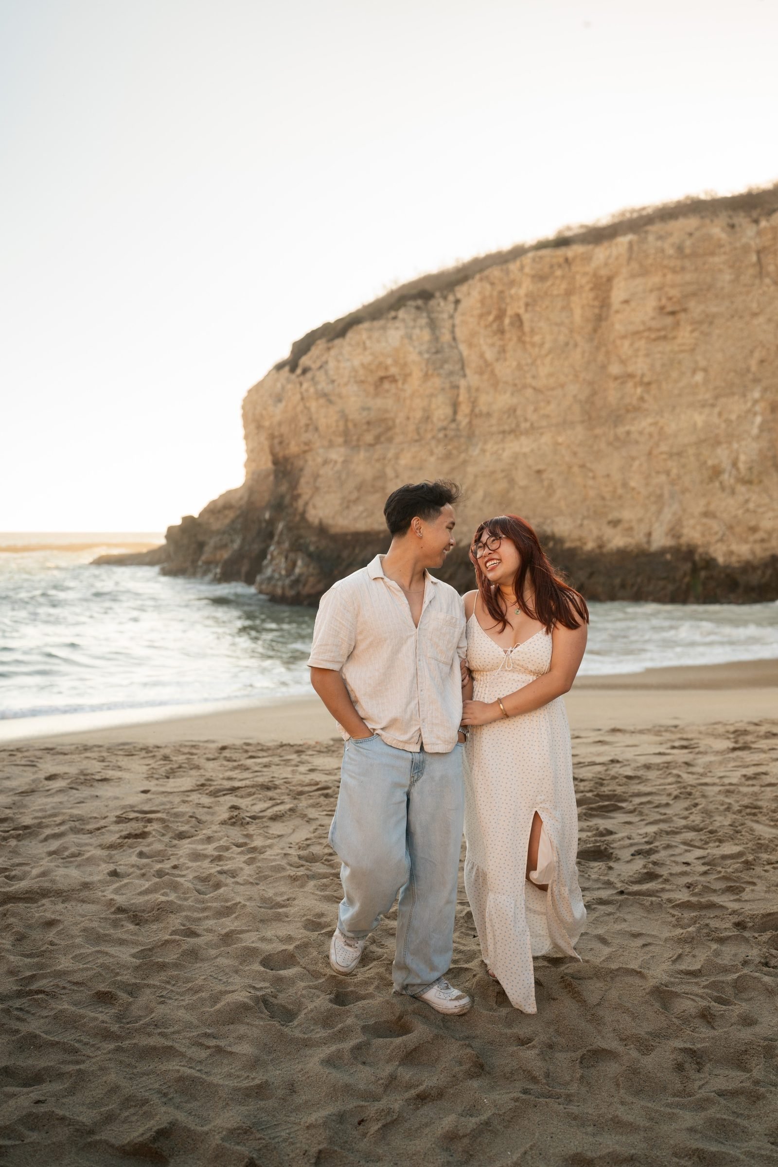 A couple laughs and leans into each other on the sandy beach at Shark Fin Cove during golden hour, dressed in coordinated neutral earth tones — a white linen shirt and light jeans, and a flowy white floral maxi dress — with a dramatic sandstone cliff and surf behind them.