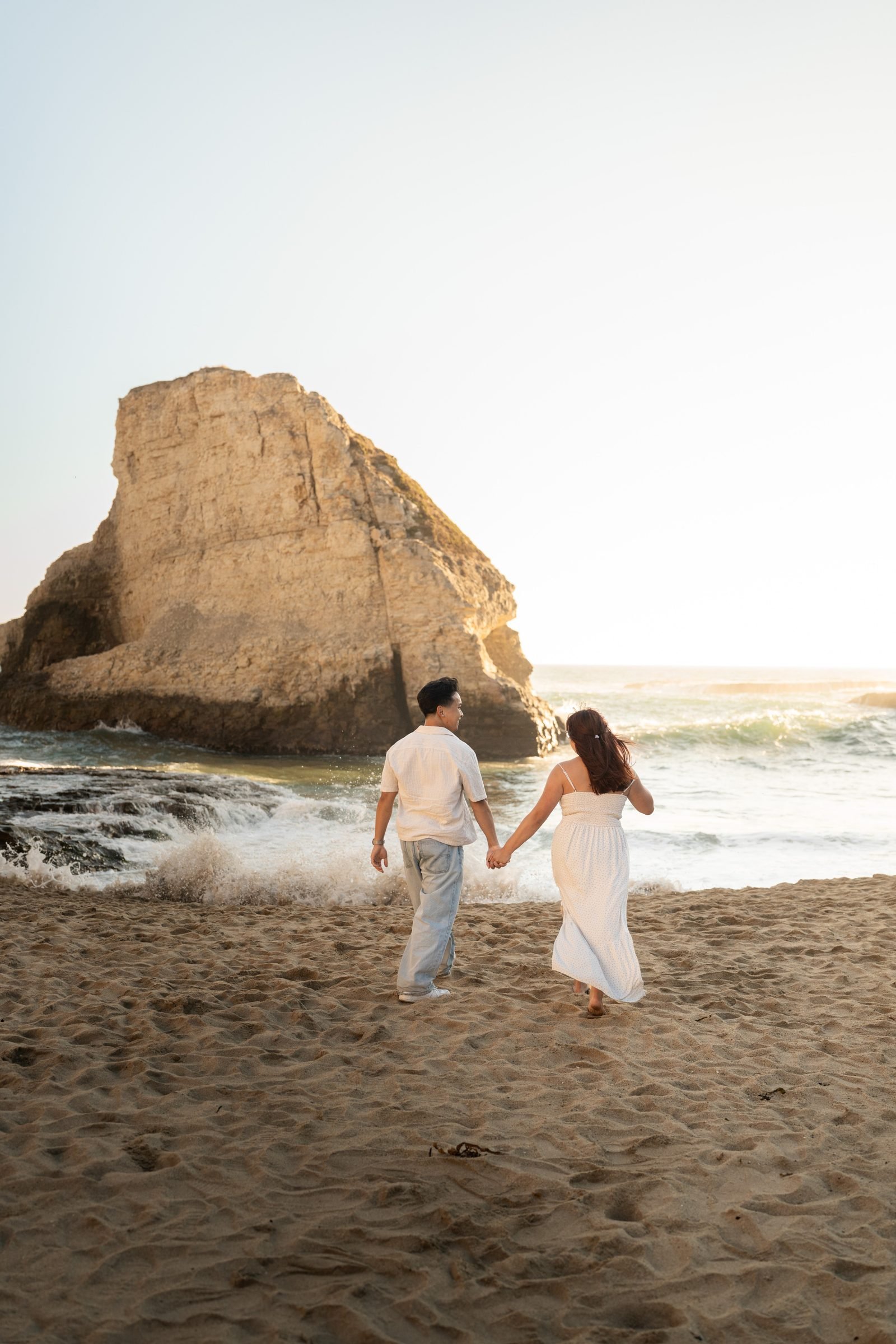 A couple walks hand-in-hand toward the ocean at Shark Fin Cove during golden hour, both dressed in white and light neutrals, with the iconic sea stack glowing warm gold behind them.